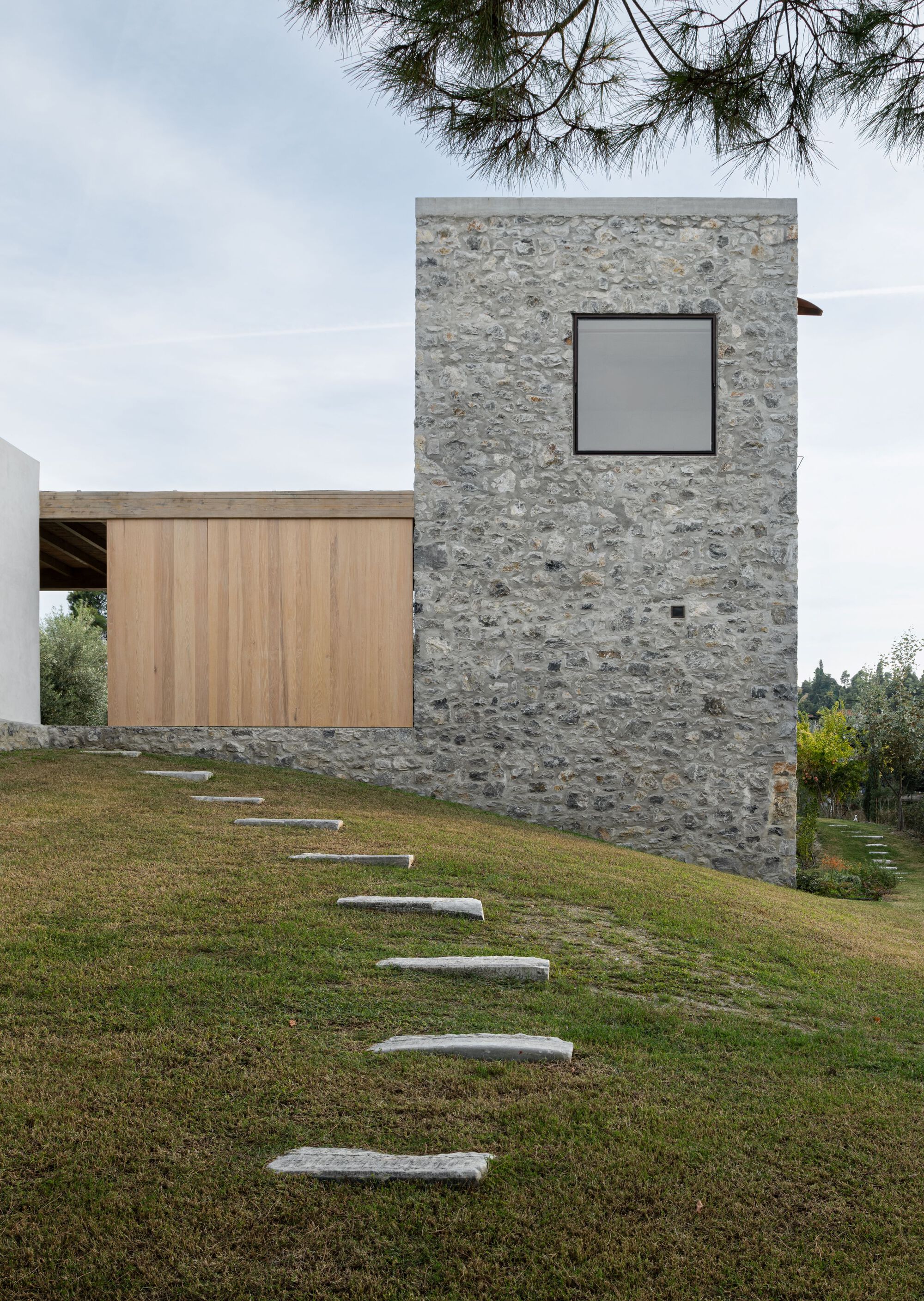 Stone tower with a square window beside a timber-clad volume, with stepping stones climbing a grassy slope under a pine branch