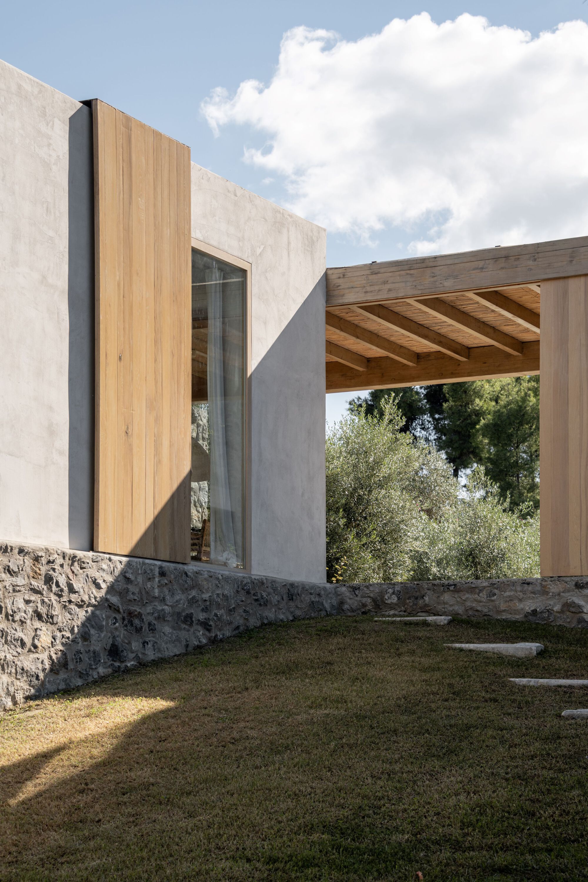 Plastered wall with a tall timber shutter and glazed opening beside a timber pergola, with hard shadows falling across the lawn