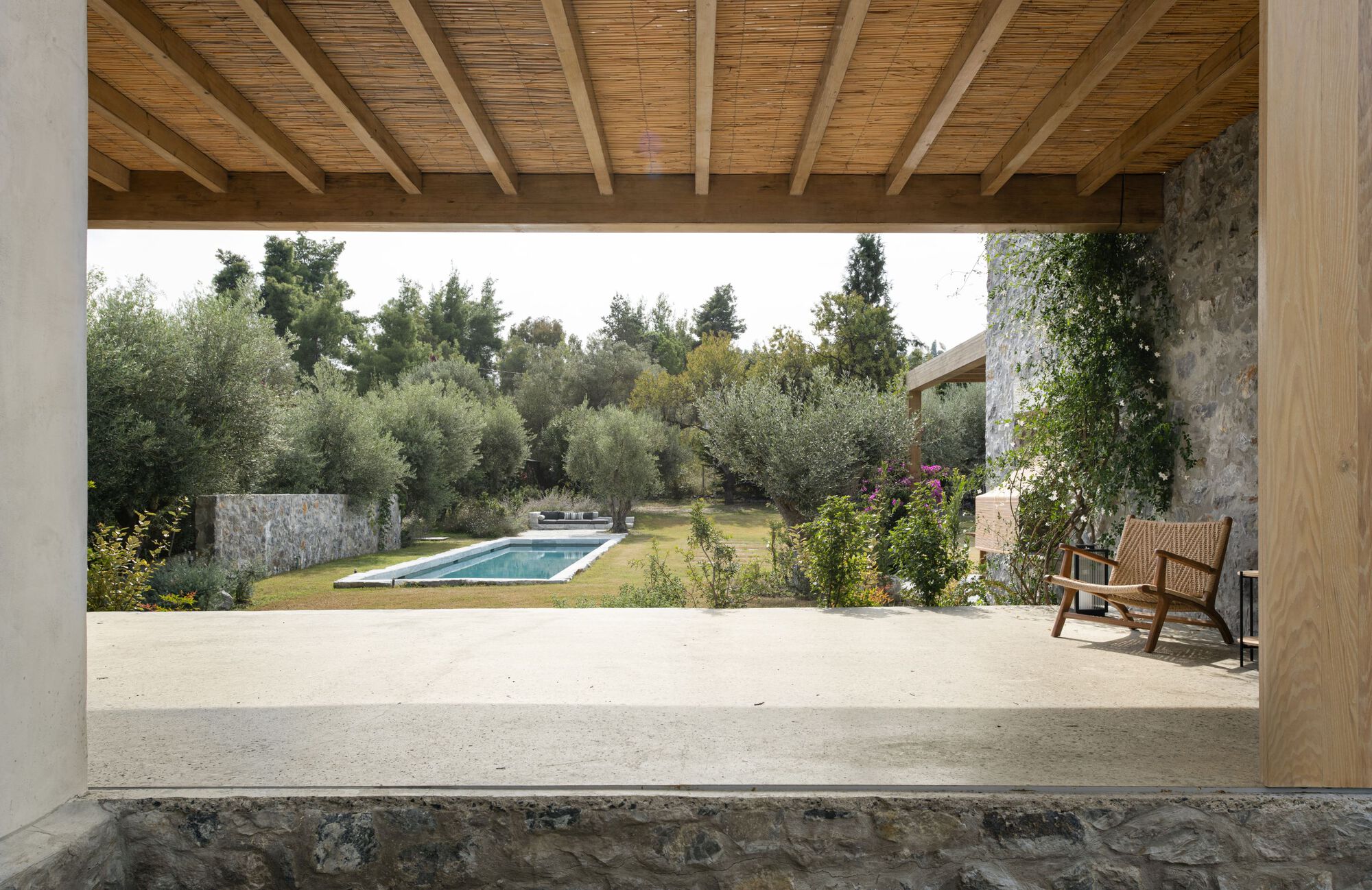 View from a covered terrace with reed ceiling and timber beams across a lawn and pool, with a wicker chair among olive trees