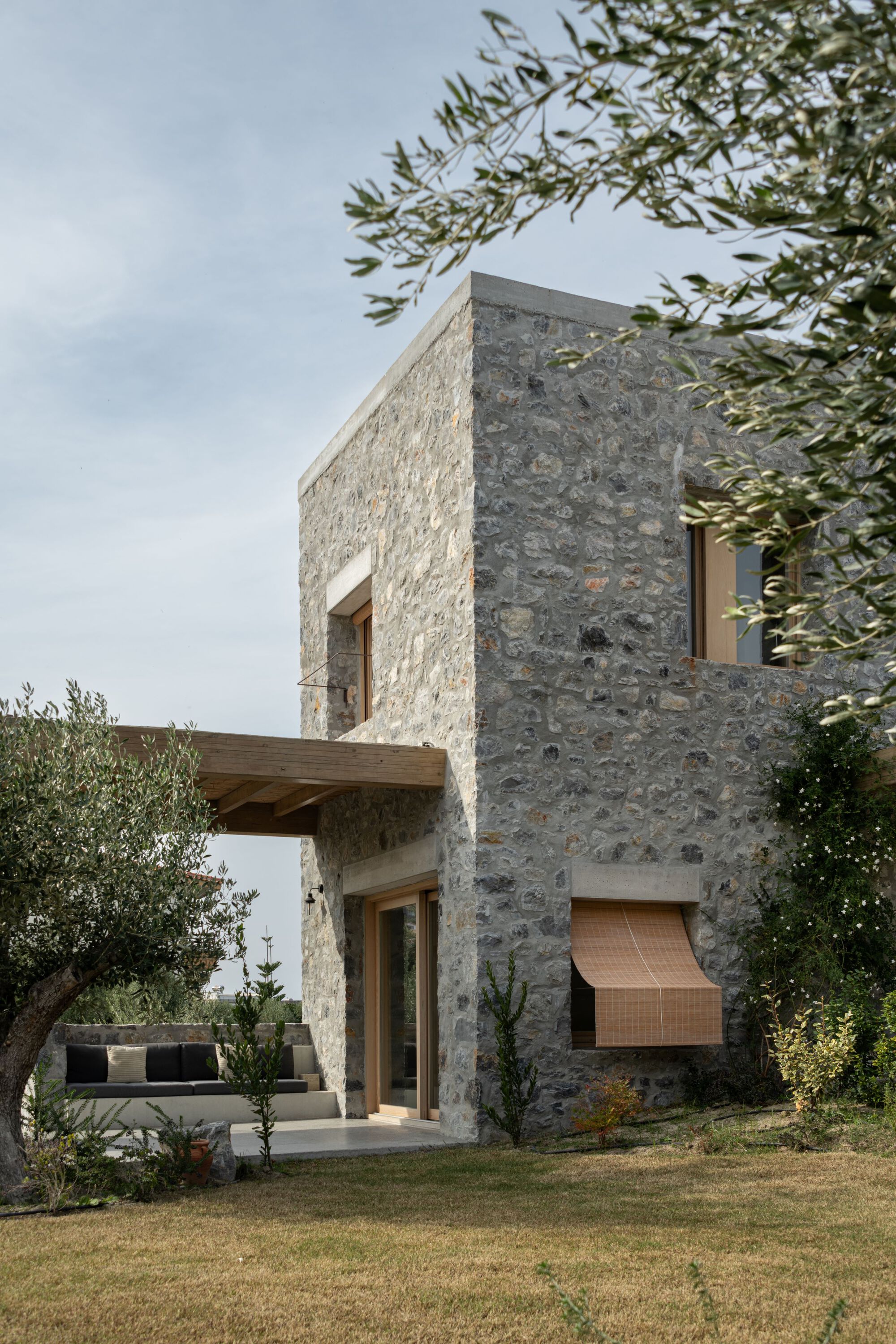 Two-storey stone house with a timber canopy over a built-in daybed, a woven blind and an olive branch against a pale sky