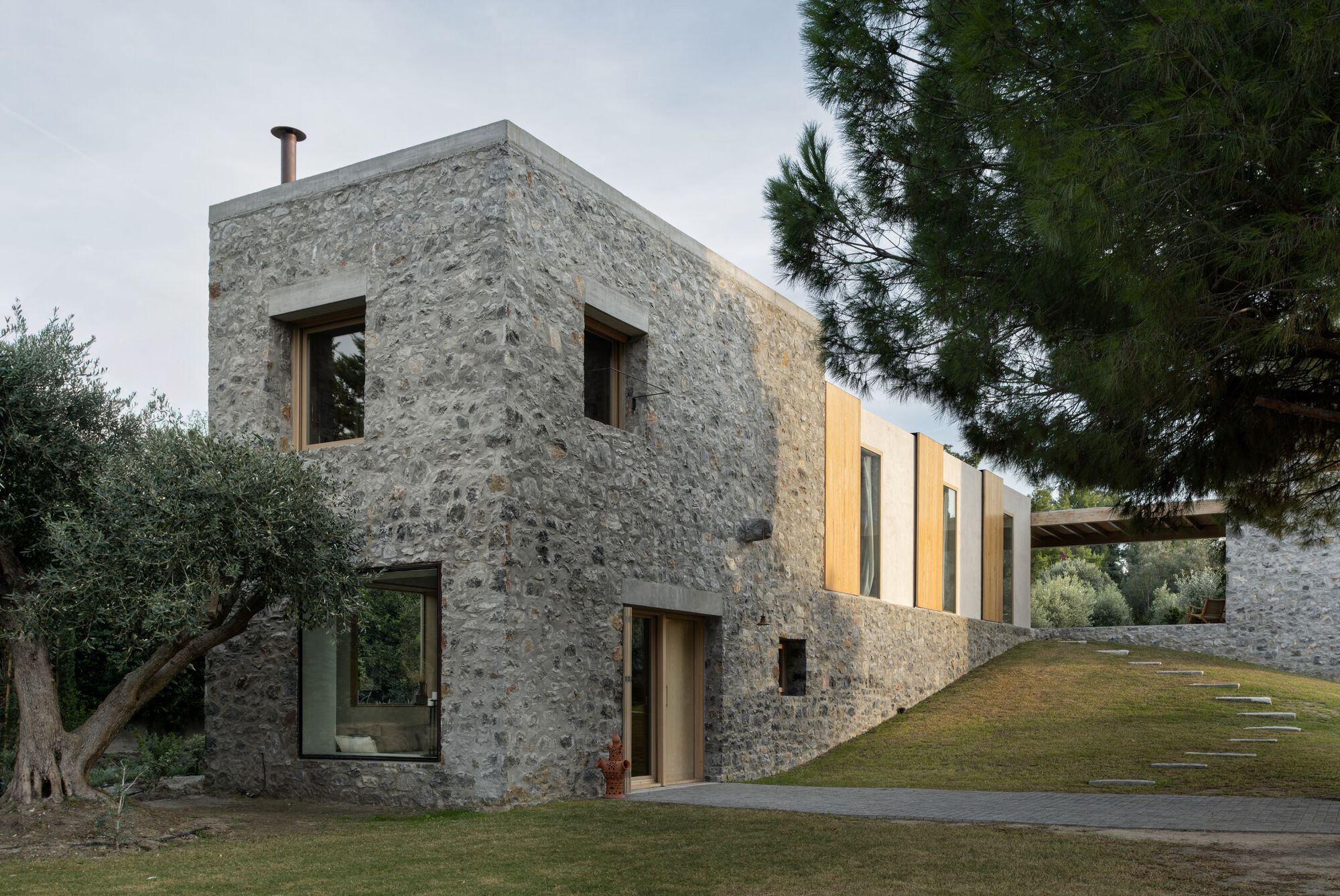 Two-storey rubble stone volume with timber-framed windows and a steel flue, beside an olive tree and a grassy slope at dusk