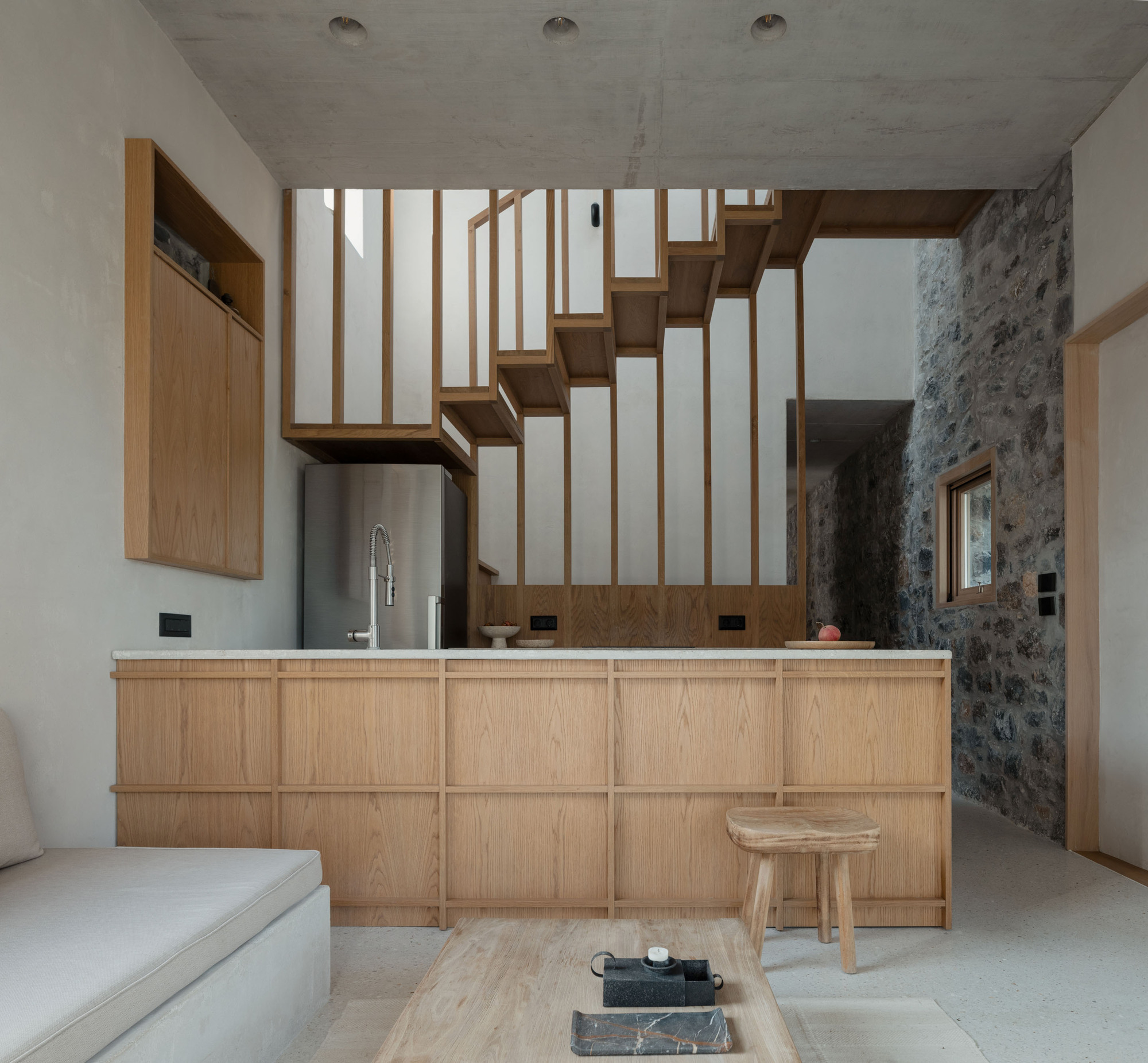 Oak-panelled kitchen counter below a floating timber stair with slatted screen, set against a rough stone wall and concrete ceiling