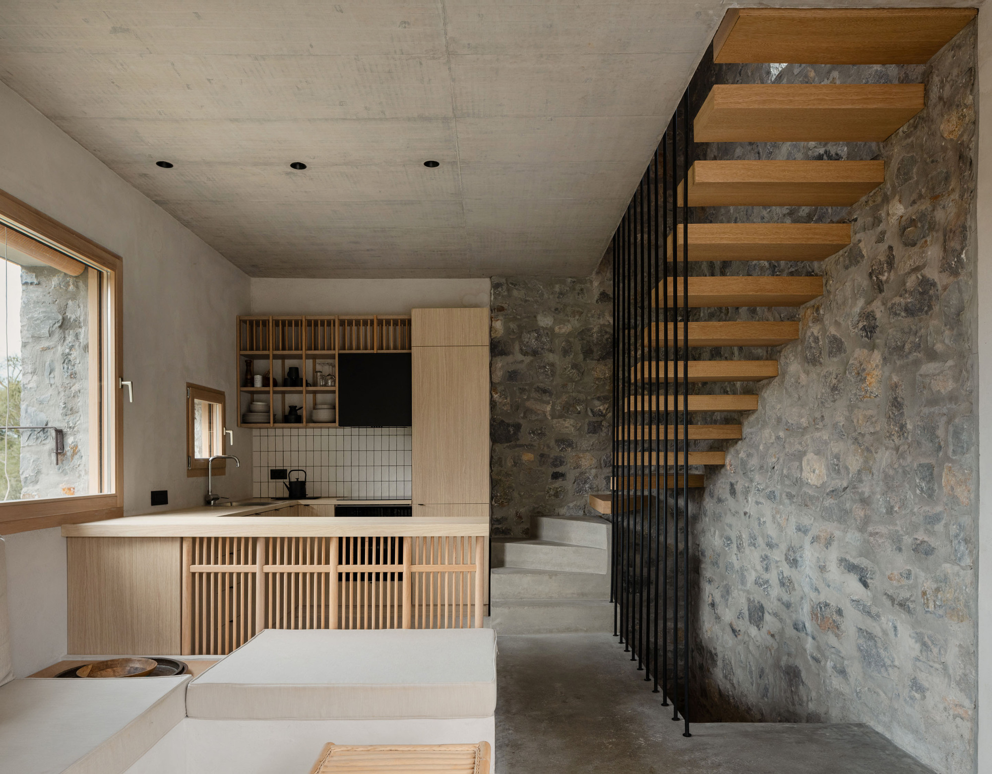 Cantilevered oak treads climbing a rough stone wall beside black steel balusters, with a timber kitchen and slatted counter beyond