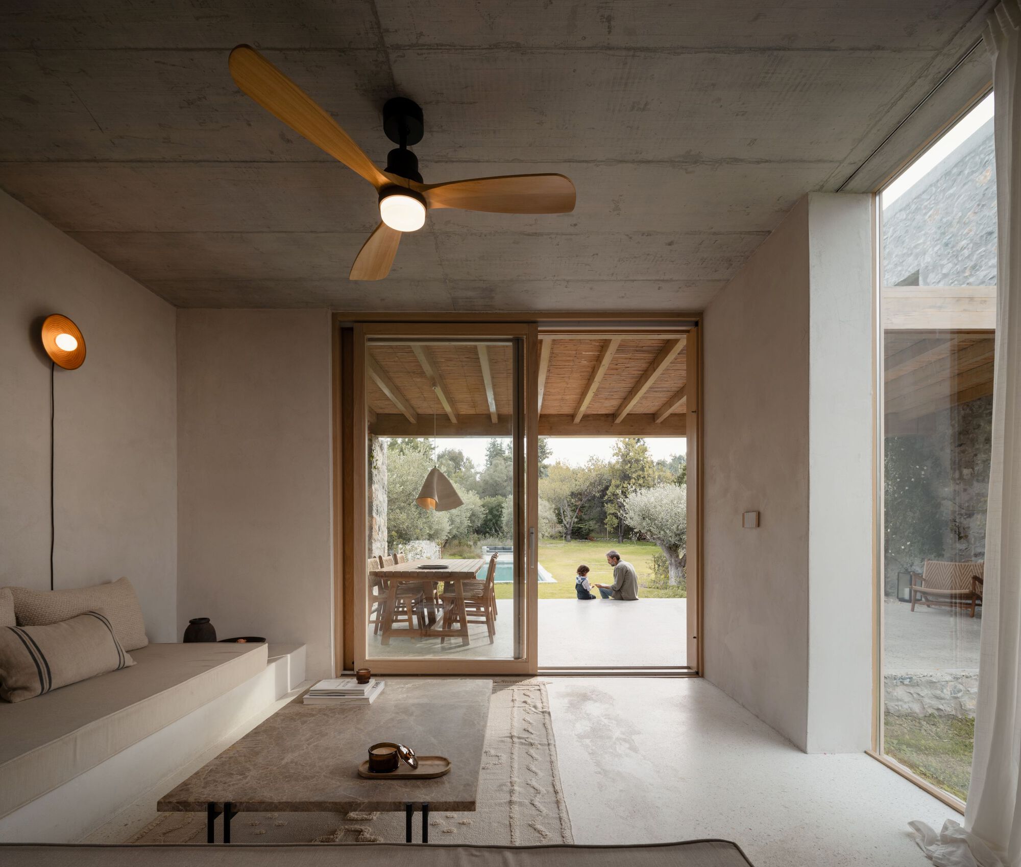 Living room with built-in sofa and ceiling fan opening through sliding glass to a covered terrace where a man and child sit on the lawn