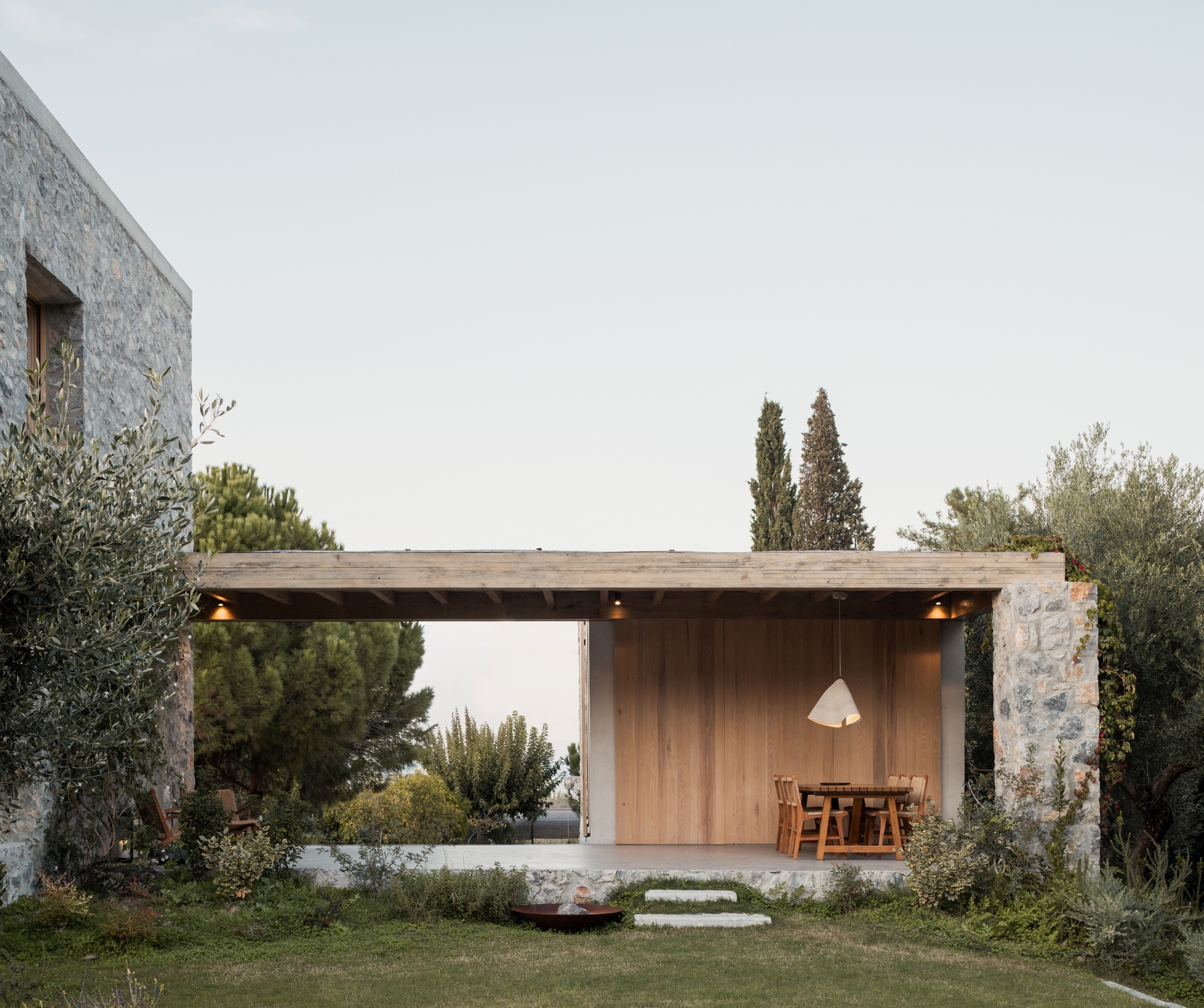 Open timber-roofed pavilion with oak-panelled back wall and dining table, framed by a stone pier, cypress trees and olive branches at dusk