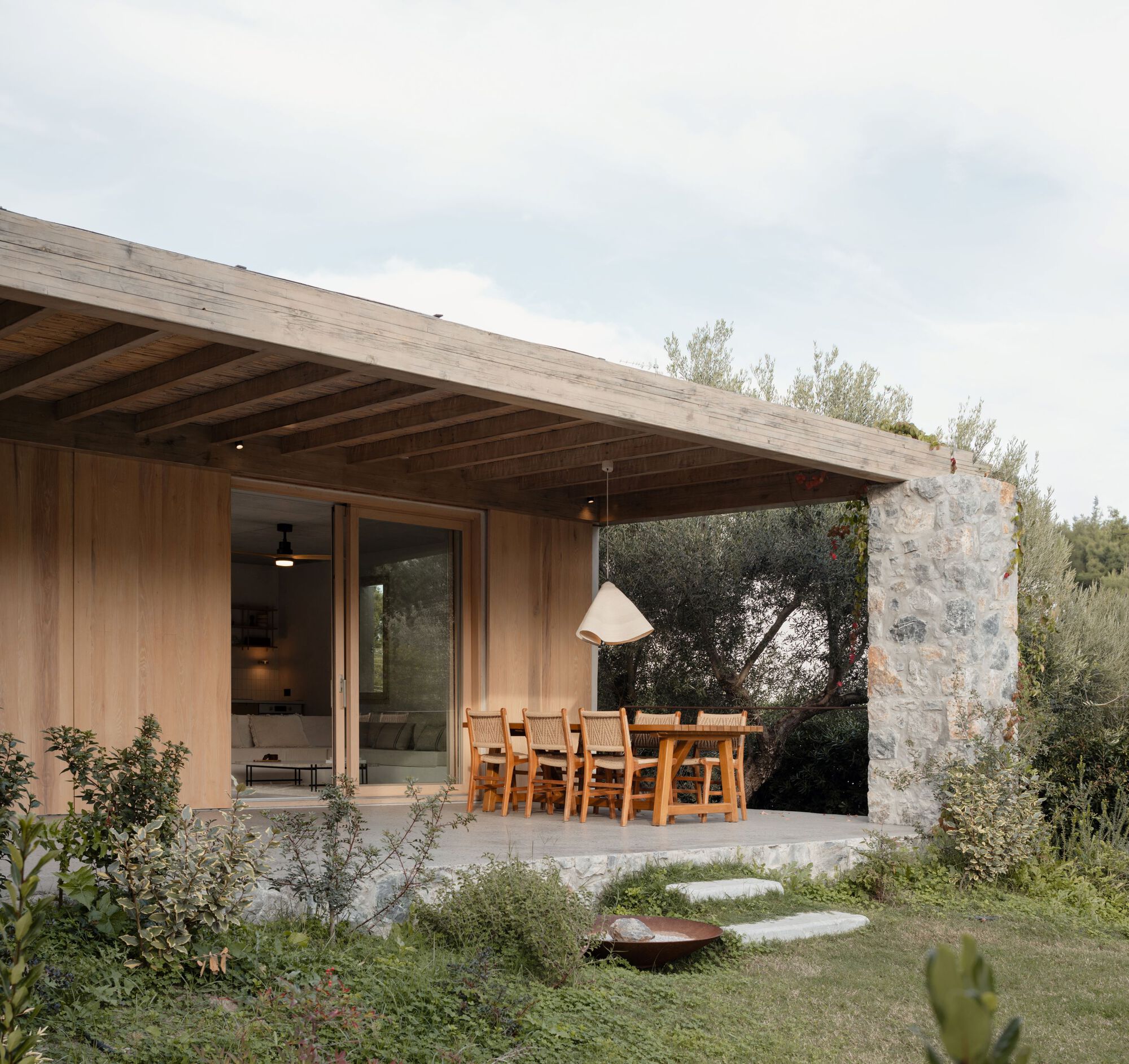 Covered terrace with timber beams, woven dining chairs and a pendant lamp beside a stone pier, under soft overcast sky