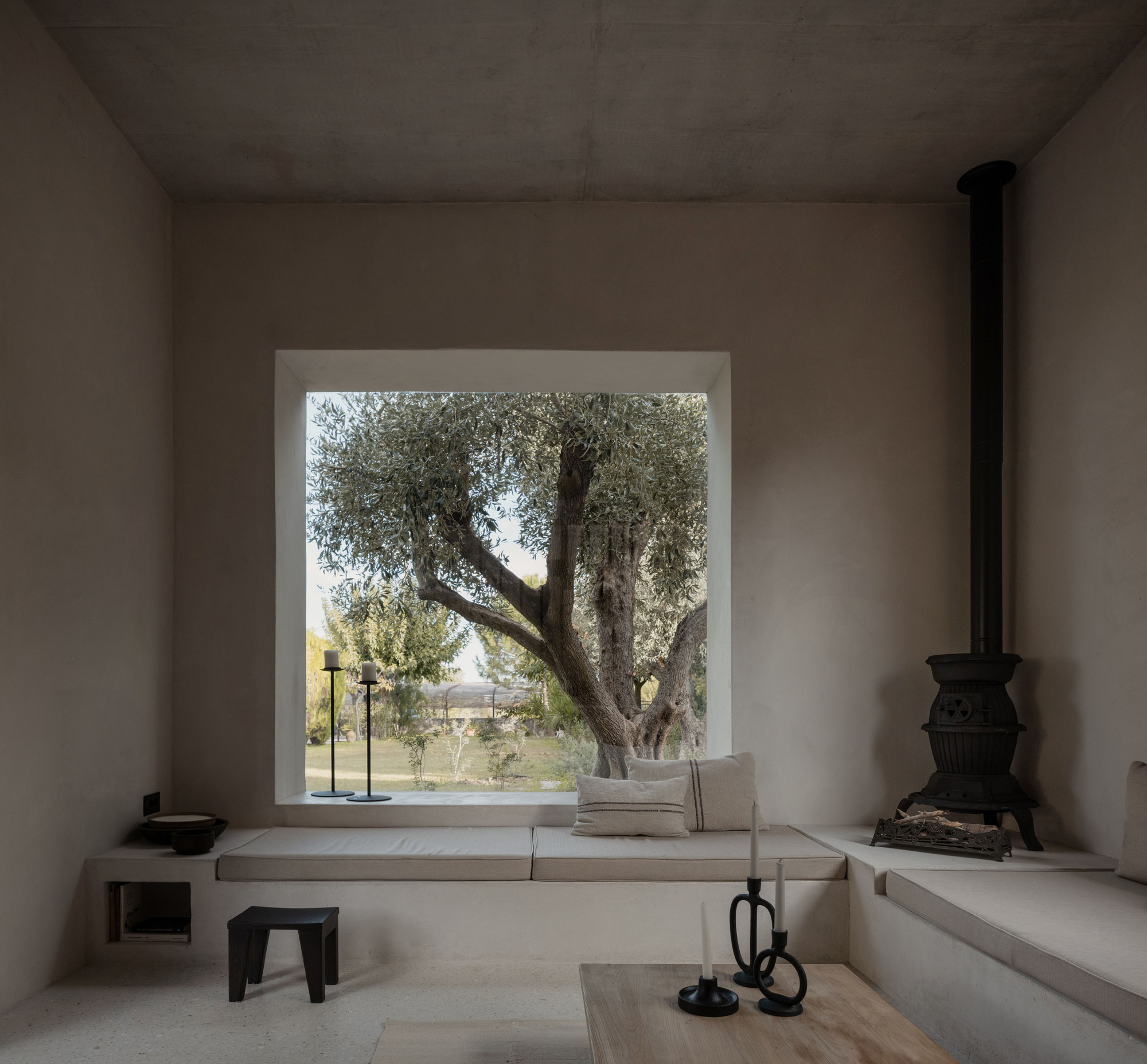Living room with a square window framing an olive tree, a built-in bench and a black cast-iron stove