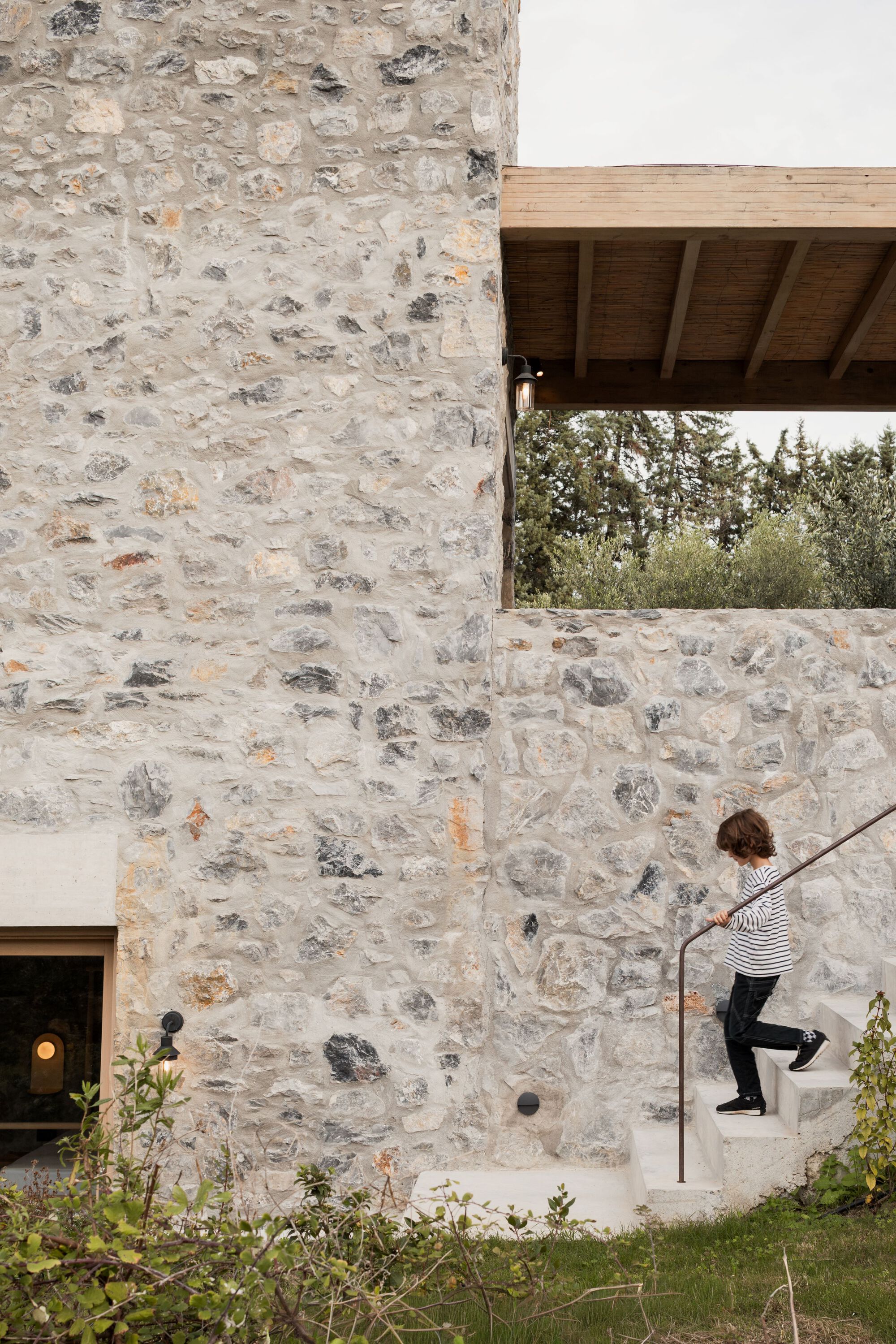 Child walking down concrete steps beside a rubble stone wall and a timber-roofed overhang, with a steel handrail