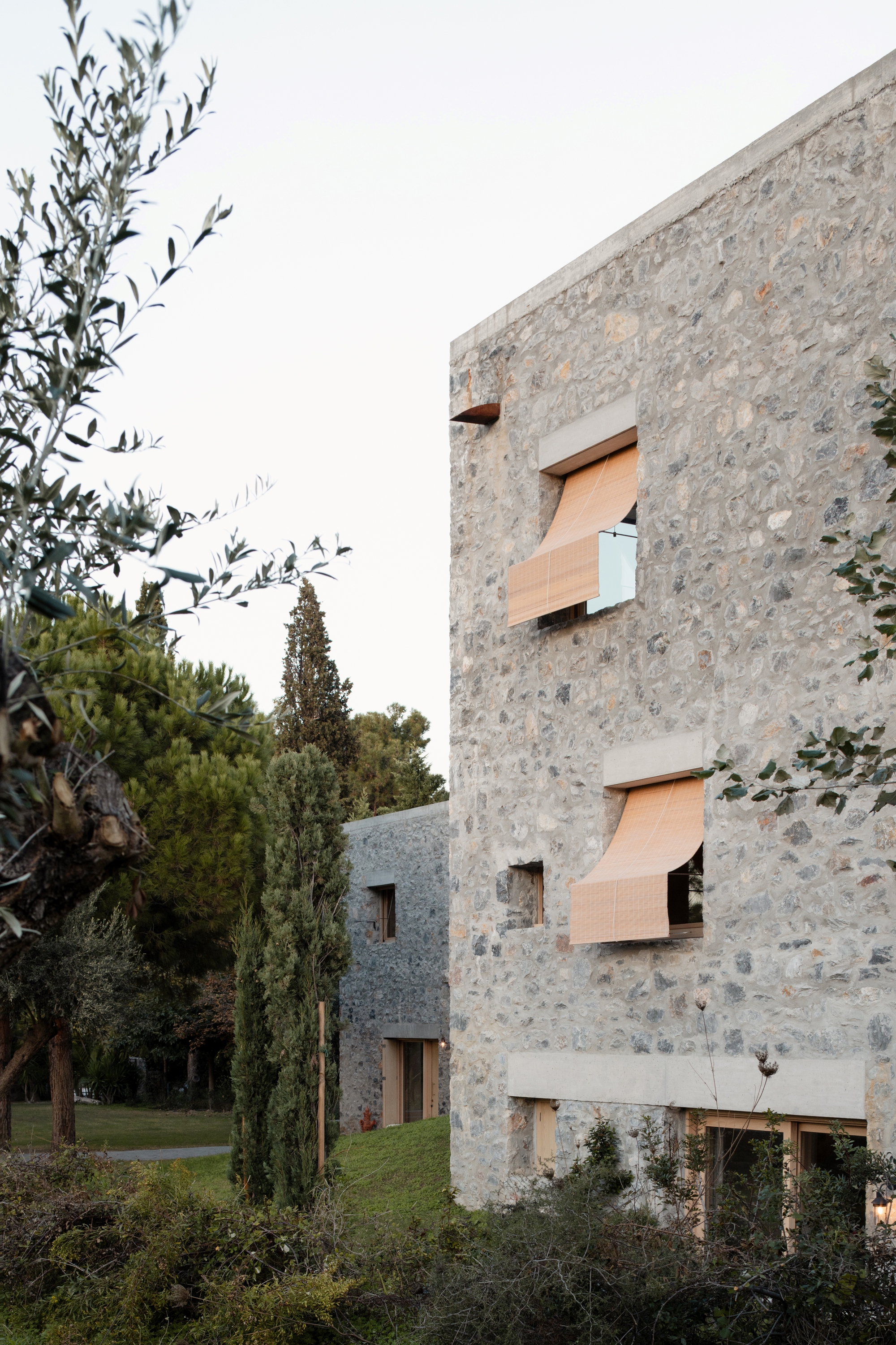Stone facade with timber-slatted awnings over square windows, seen past olive and cypress trees in soft evening light