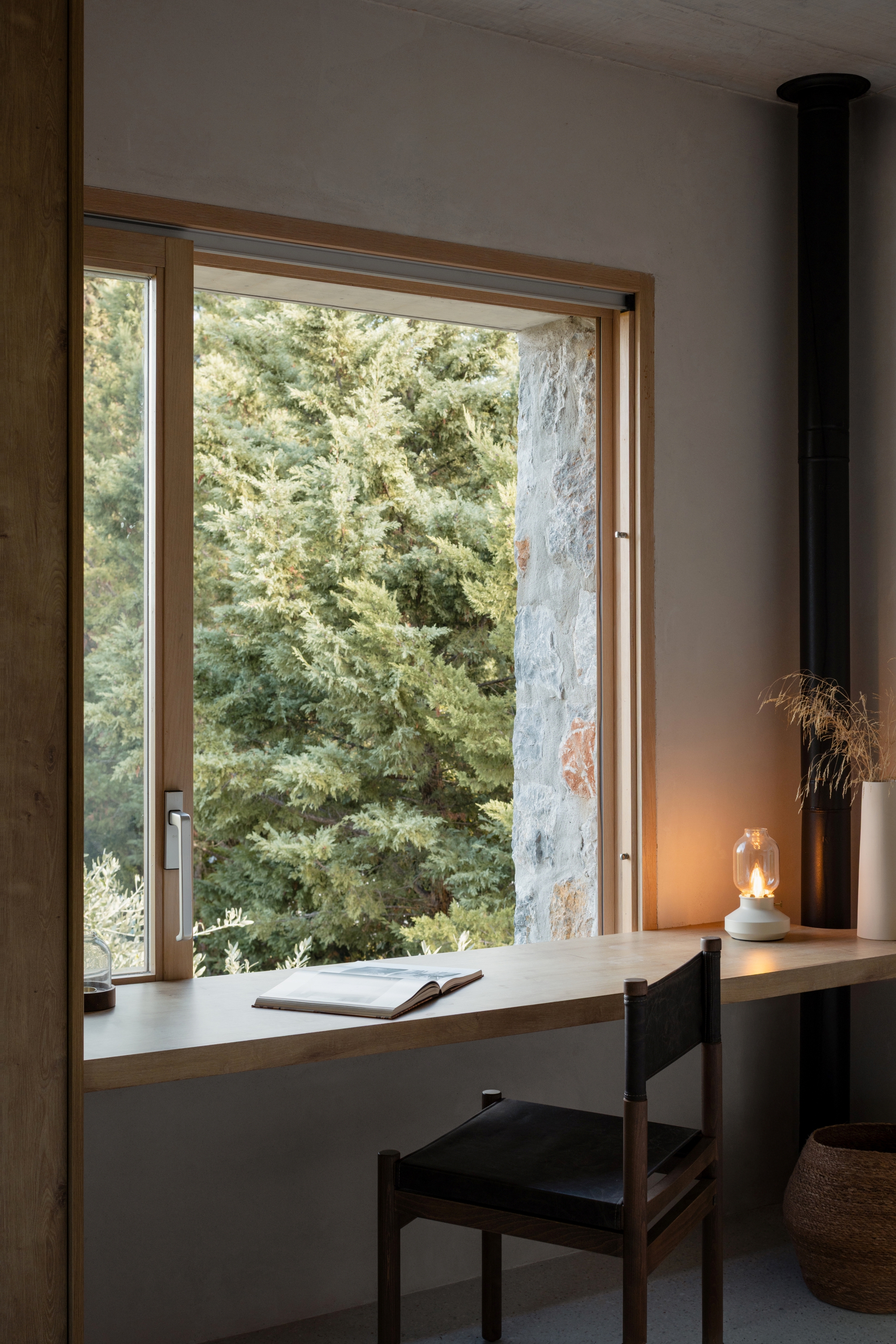 Timber desk running along a corner window with an open book, a lit lamp and a black stove flue