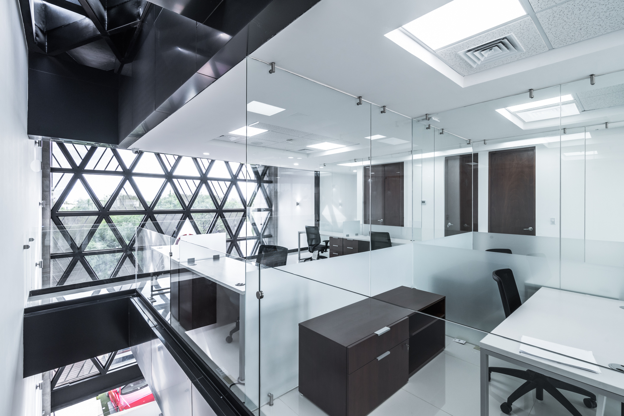 Open office floor behind frameless glass partitions, with the triangulated facade grid and a red car visible below through the window