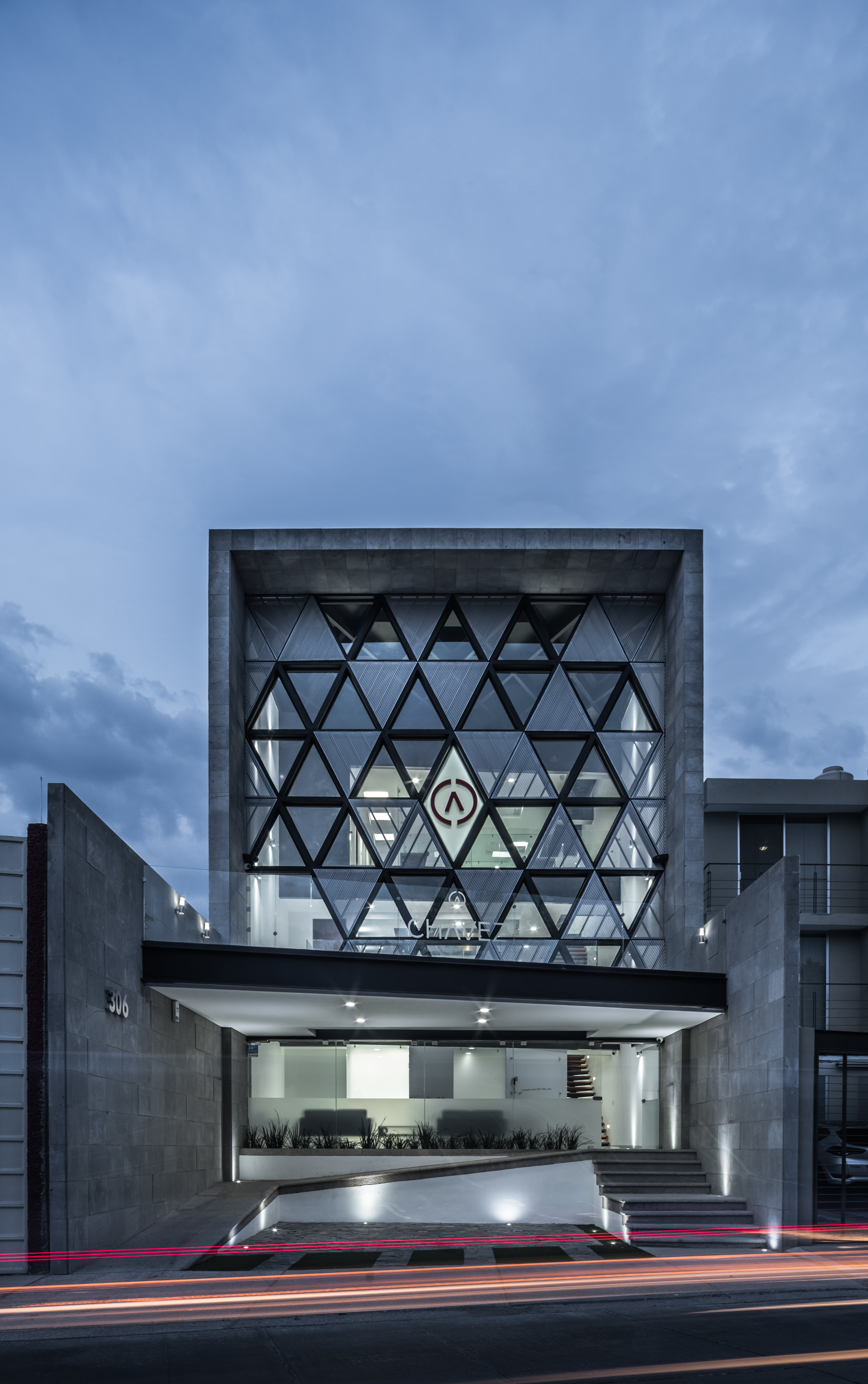 Front elevation with a triangulated glass facade framed by a stone box, seen at dusk with red light trails on the street