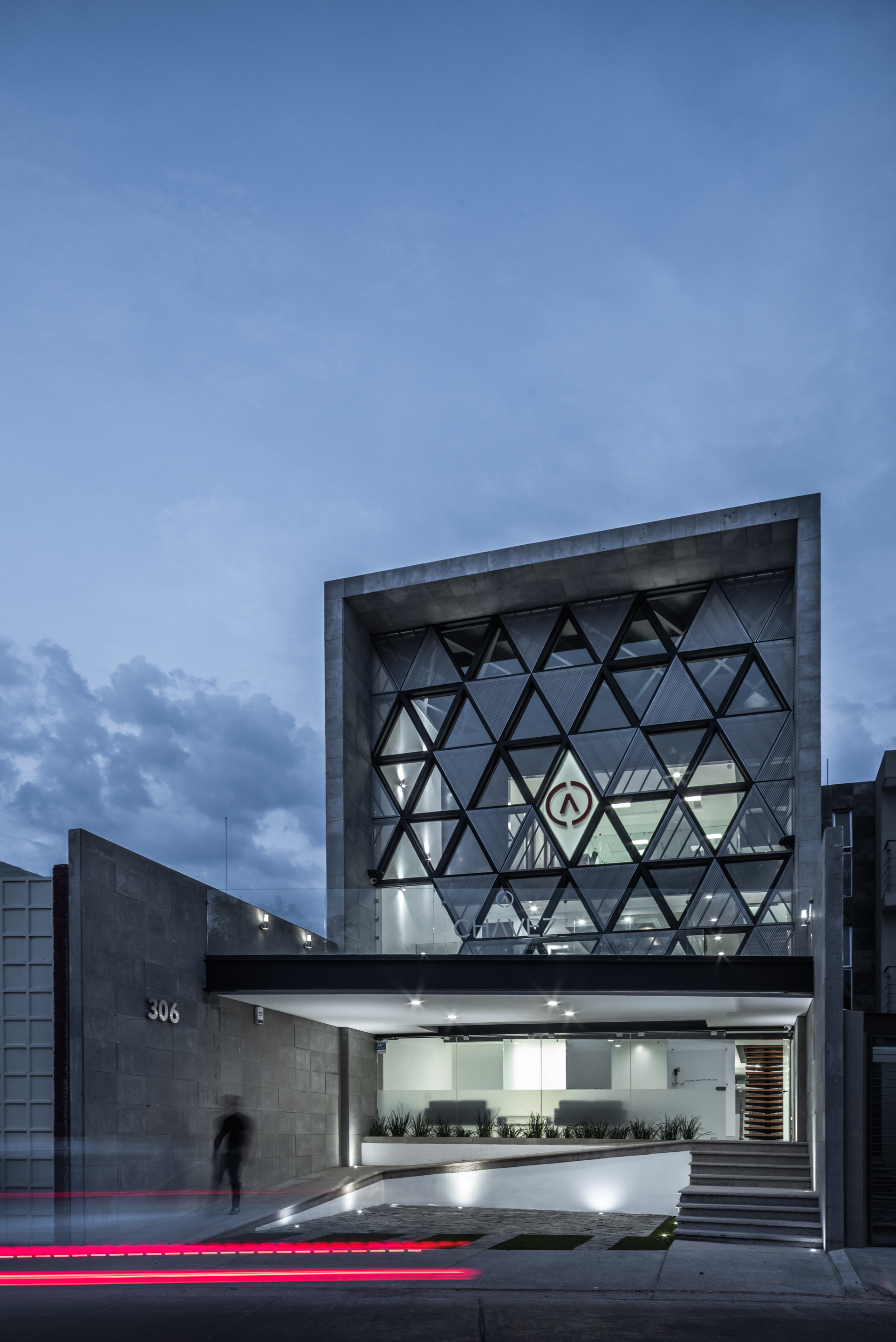 Street view of the stone-framed triangular glazing at blue hour, with a blurred pedestrian walking past the numbered wall