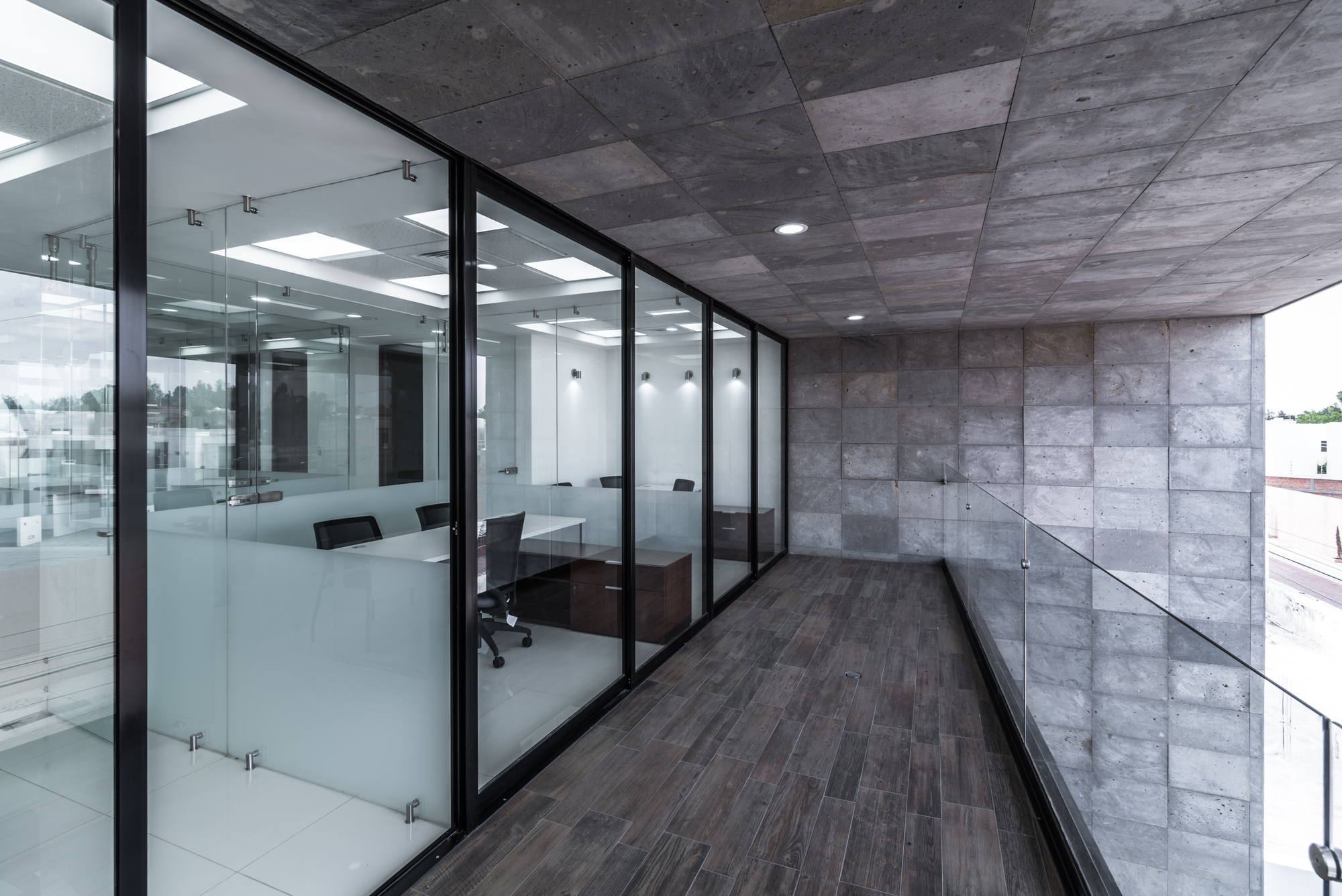 Exterior balcony walkway with stone-clad wall and ceiling, wood-look tile floor and glass-partitioned offices along one side