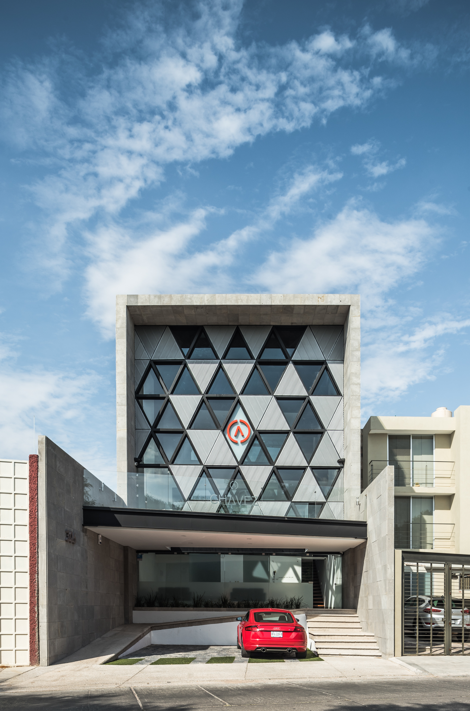 Front elevation of the triangulated glass facade under a blue sky with wispy clouds, a red car parked at the entry
