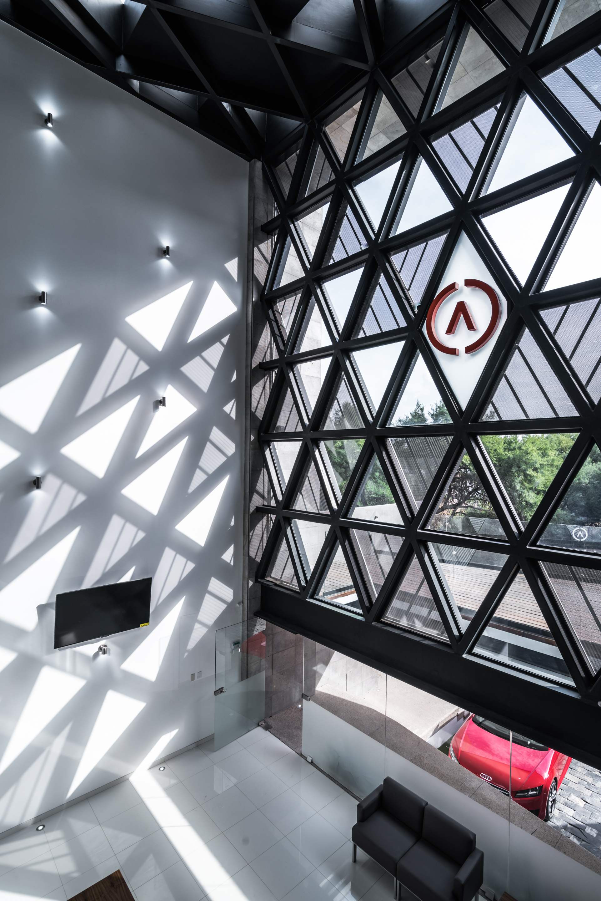 High angle view of the triangulated steel and glass facade casting sunlit triangles across a white lobby wall with a red car outside