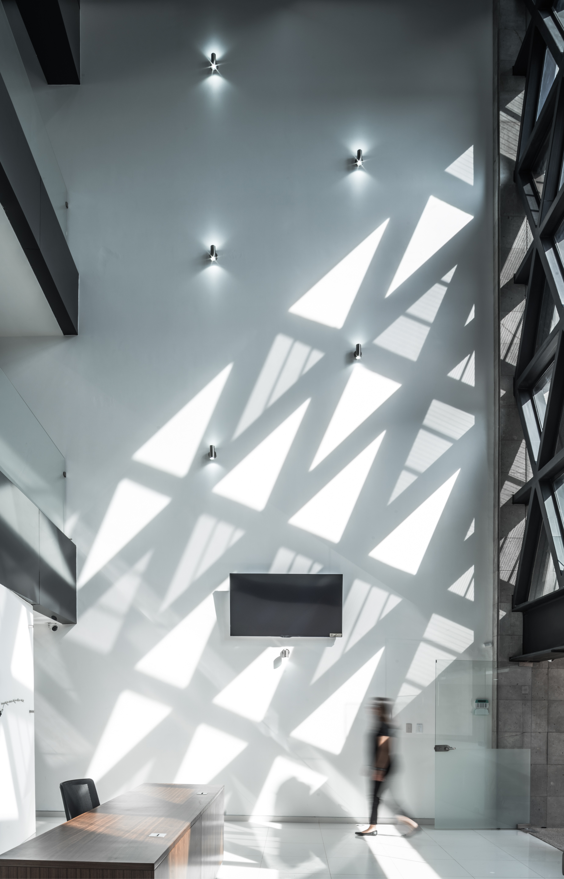 Tall white lobby wall covered in triangular sunlight patterns as a blurred figure walks past a reception desk
