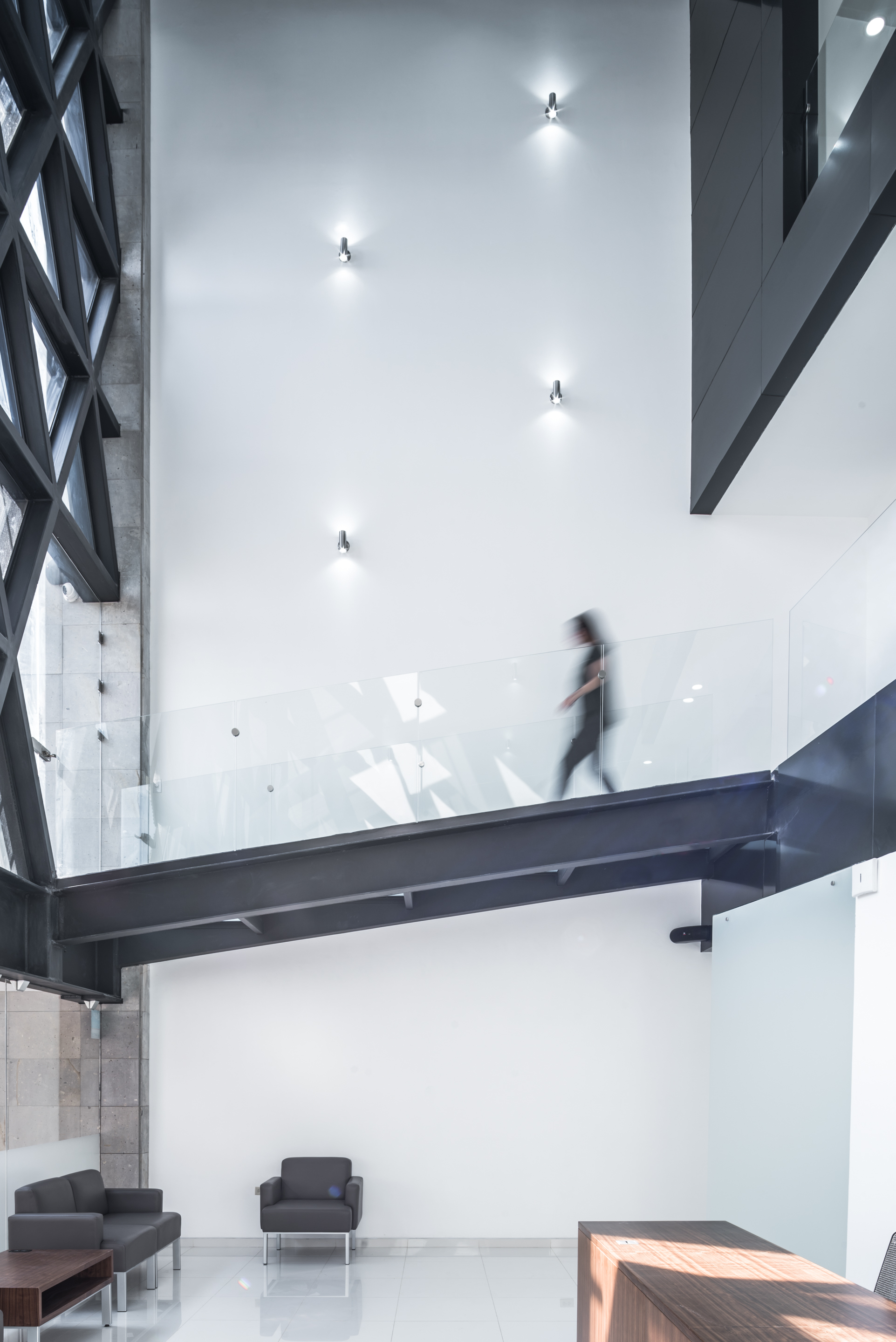 Steel bridge with glass balustrade crossing the double-height lobby as a blurred figure walks above lounge chairs