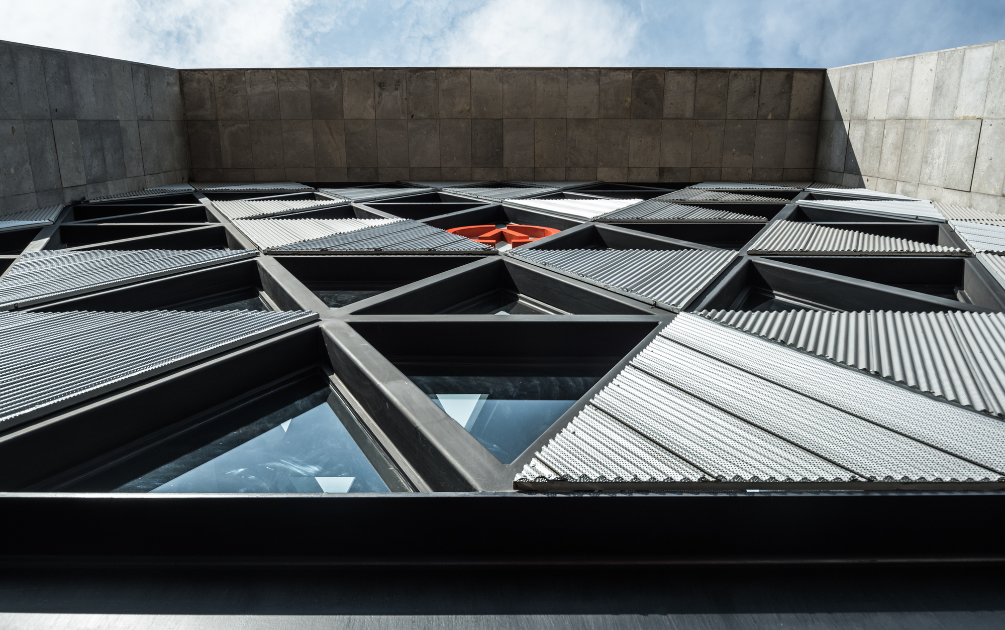 Upward view of the triangulated facade with corrugated metal panels, glazed openings and a red circular emblem under a cloudy sky