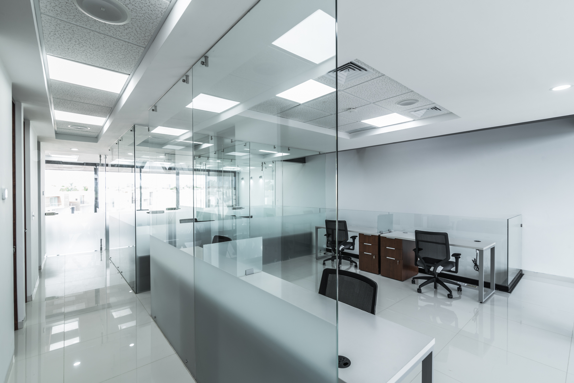Open office with frameless glass partitions, workstations and recessed ceiling lights reflected in the glossy white floor