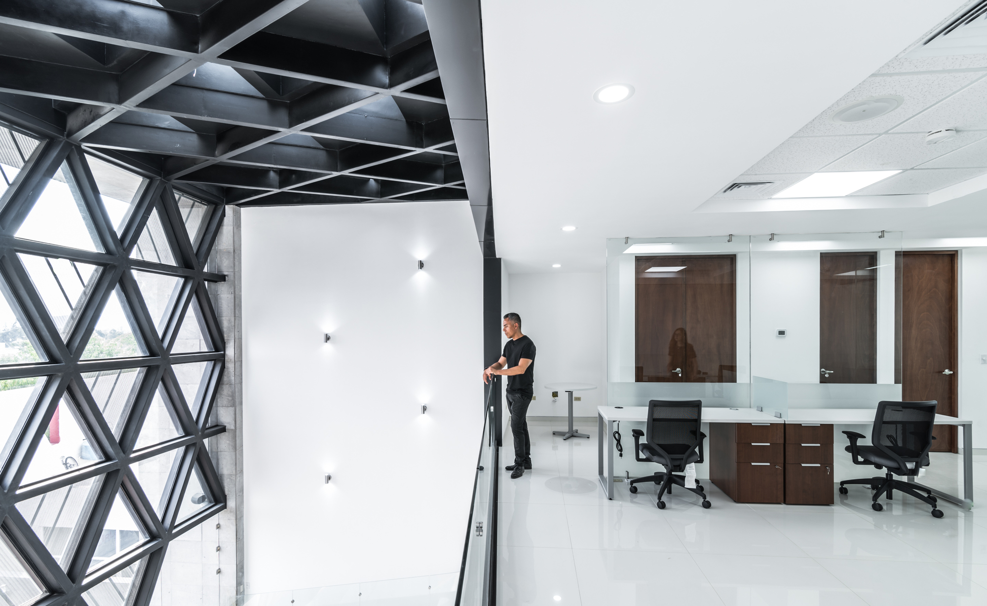Upper walkway beside the triangulated steel window wall, with a man at the glass rail overlooking office desks