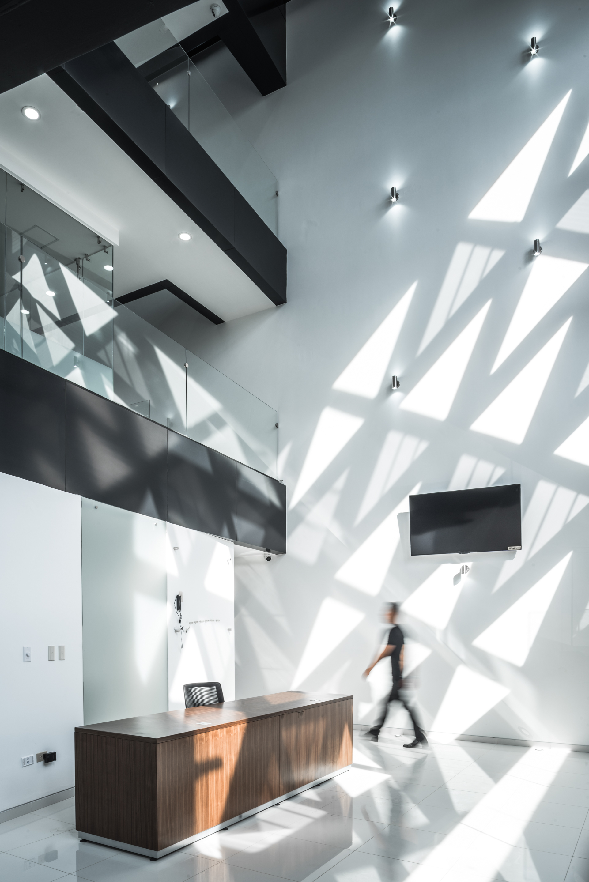 Double-height lobby with a timber reception desk and triangular sunlight patterns across the white wall as a person walks past