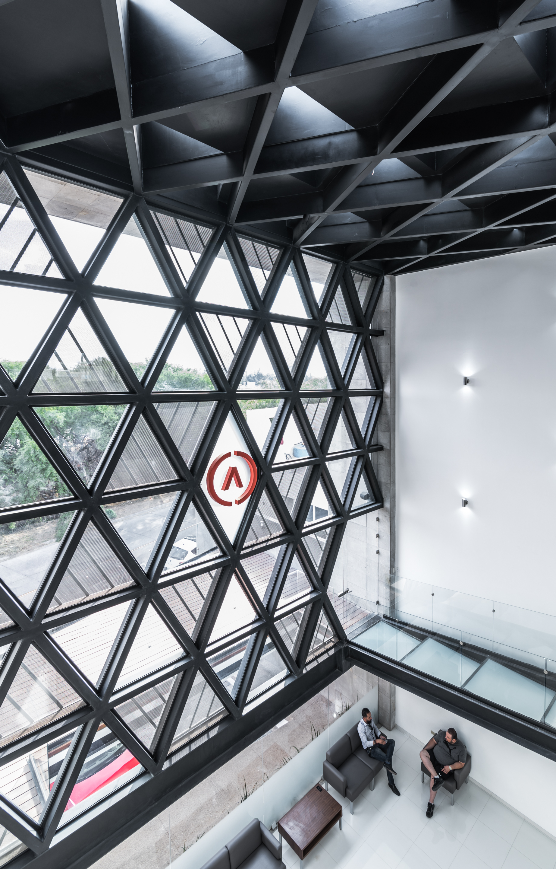 Looking down through the triangulated steel window wall toward two seated visitors and a glass-floored bridge in daylight