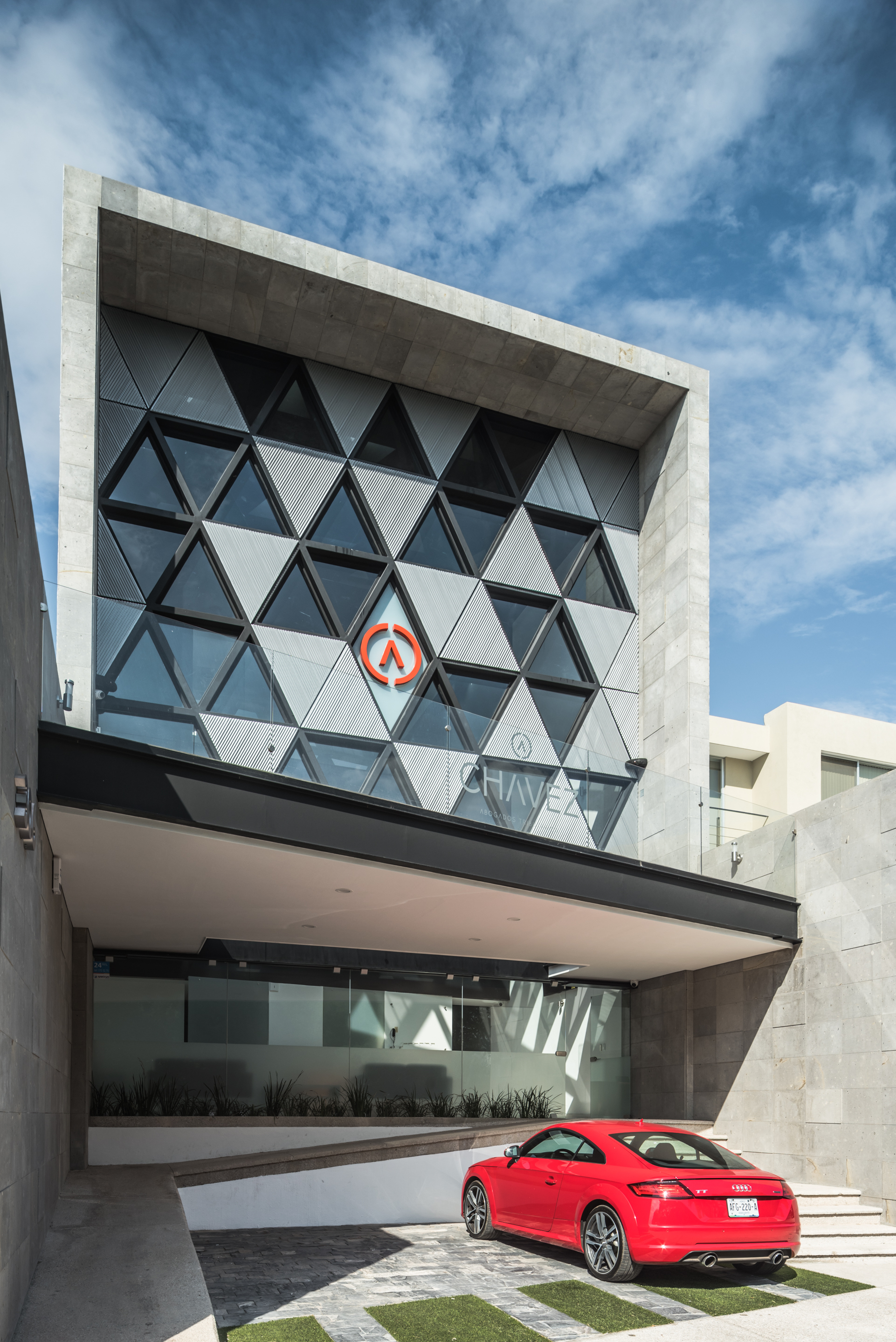 Street facade with a triangulated glass and steel grid above a deep canopy, a red car parked in front under blue sky