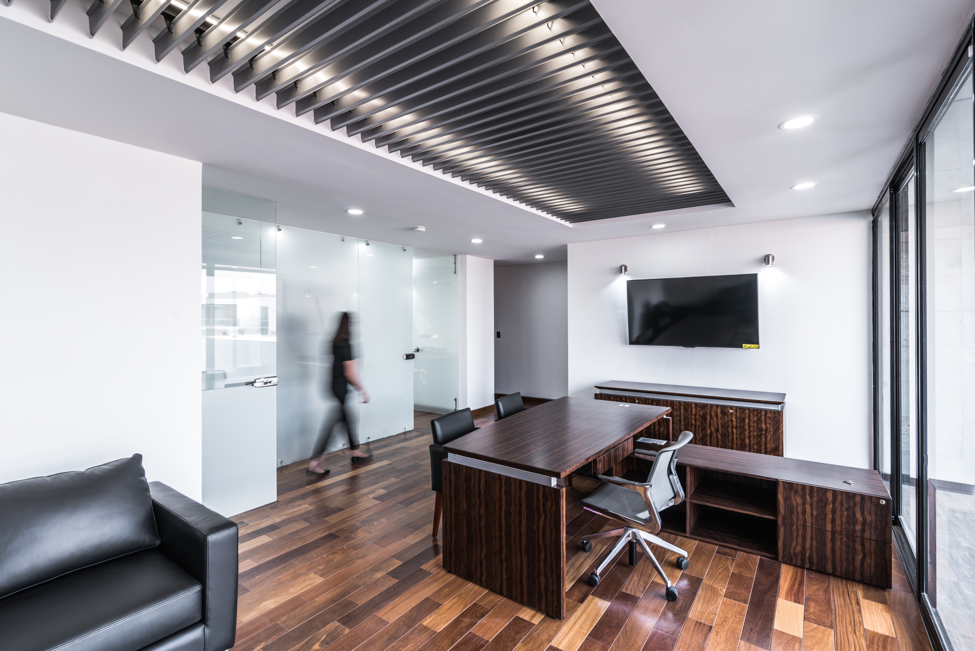 Director's office with a dark wood desk, slatted metal ceiling panel and frosted glass partition as a person walks past in blur