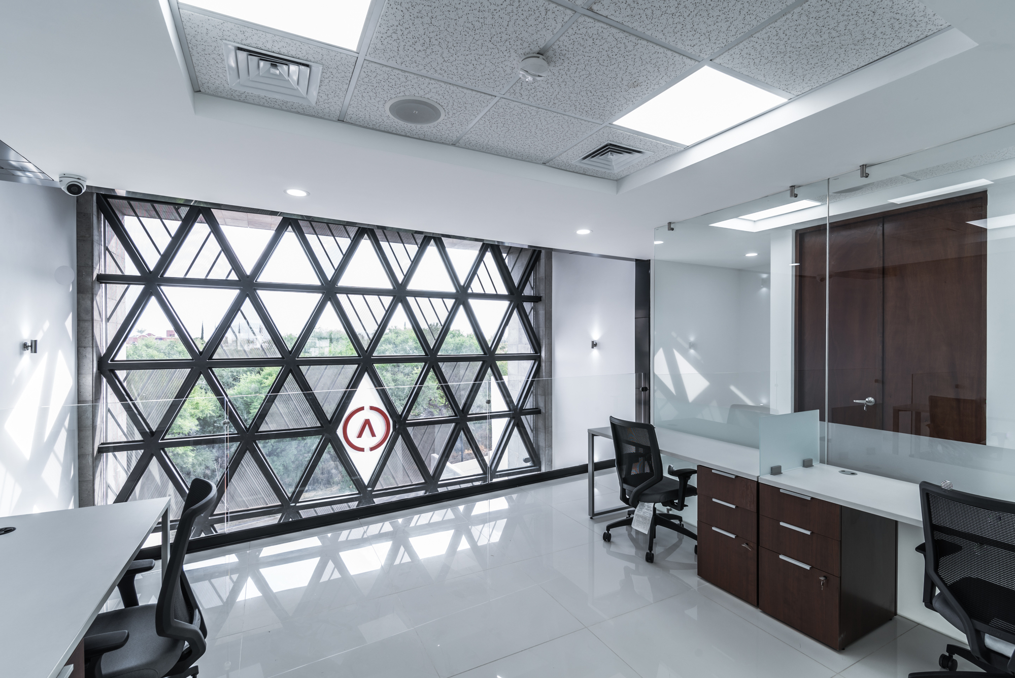 Open office beside a triangulated lattice screen over a window, with green trees visible beyond and daylight on glossy white floor tiles