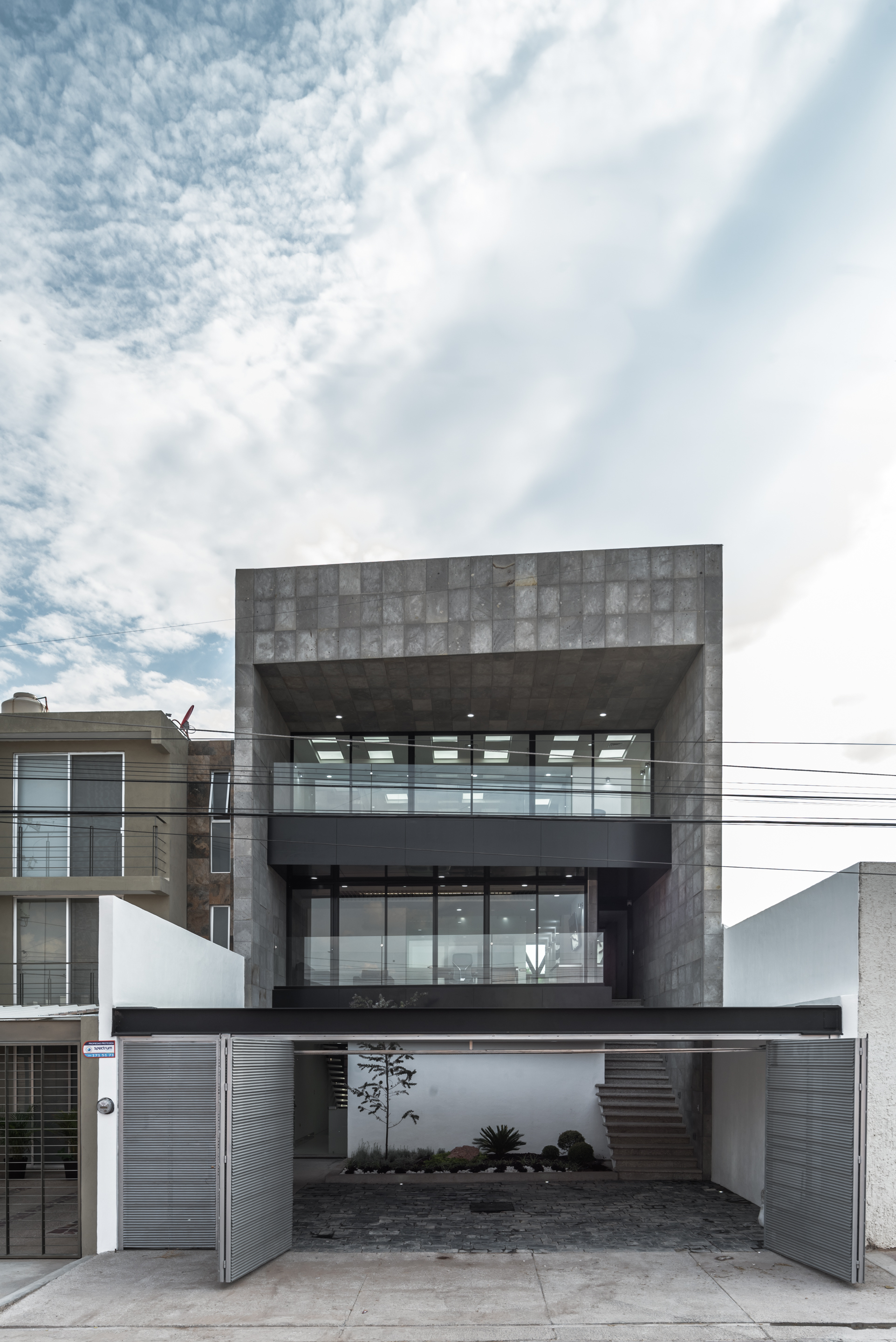 Street facade of a stone-clad upper volume above glazed floors and a folding louvered garage gate under a cloudy sky