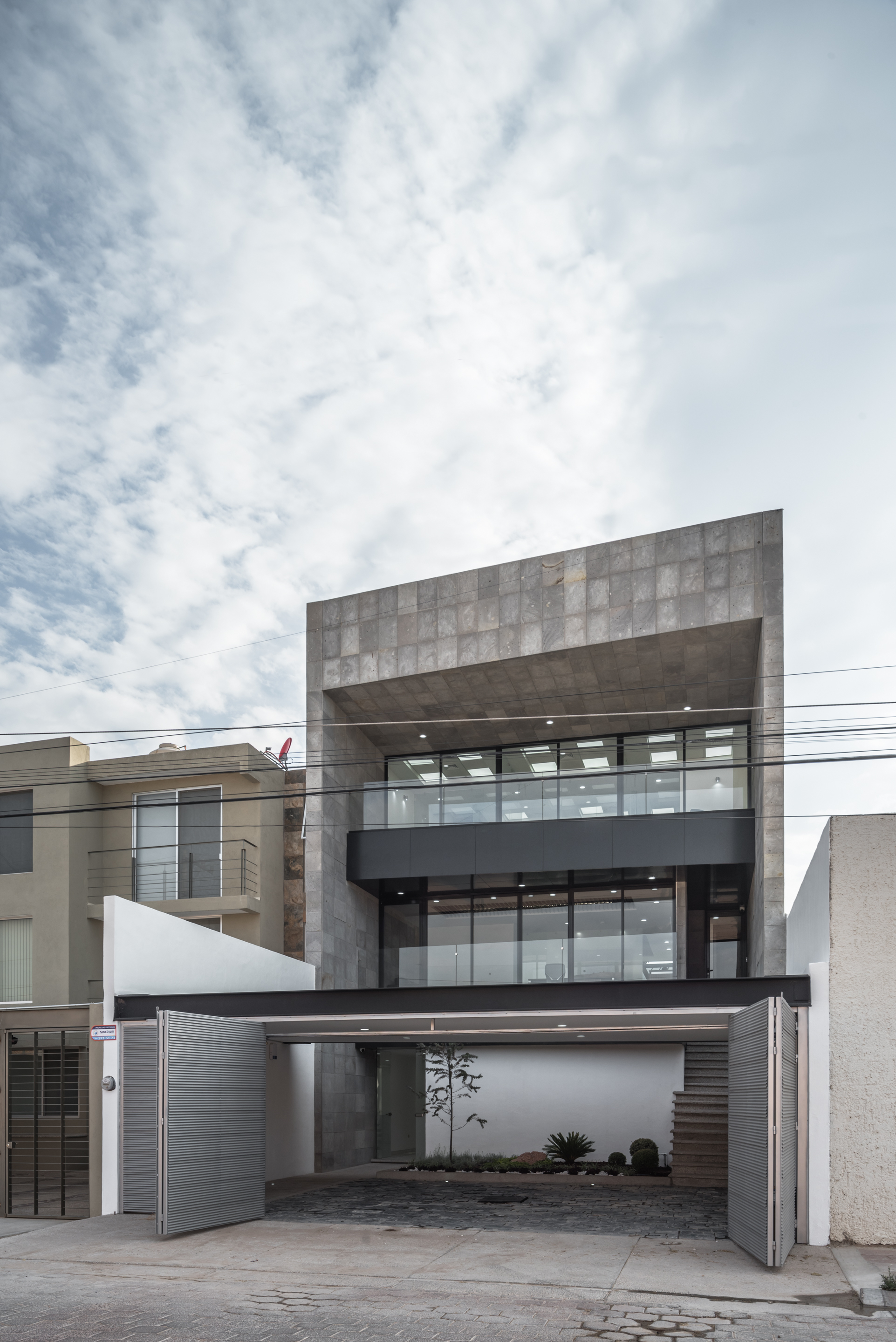 Angled view of the stone-clad street facade with a glazed office floor above an open parking court and folding gates, under thin cloud