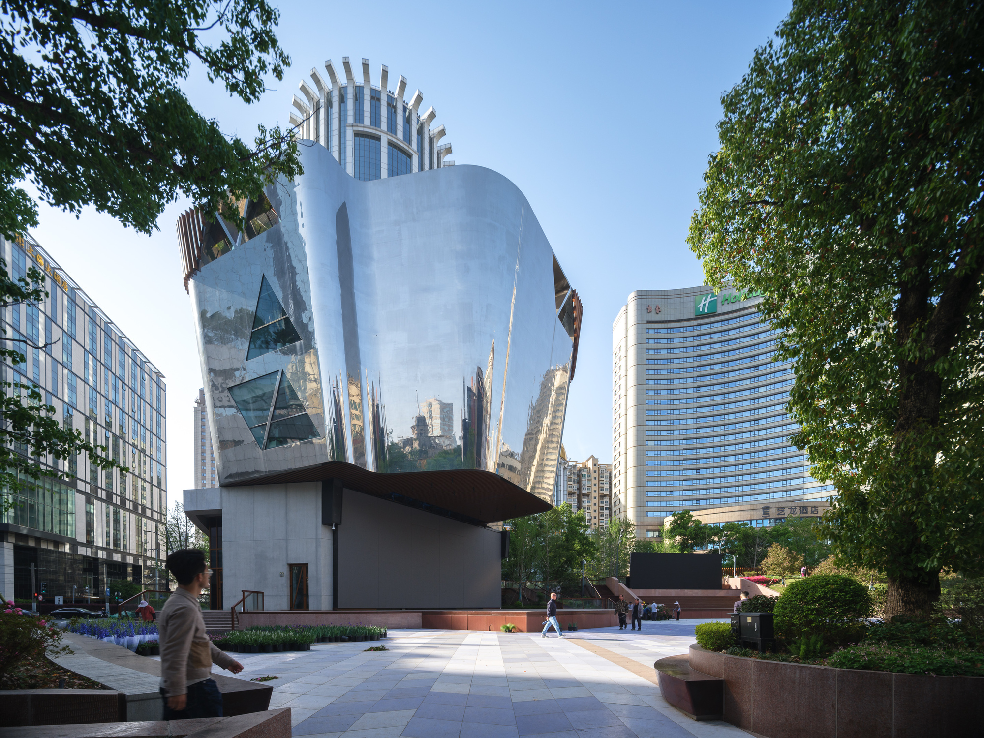 Mirror-polished steel pavilion with triangular glazed openings rising over a concrete base, seen from a paved plaza in clear afternoon light