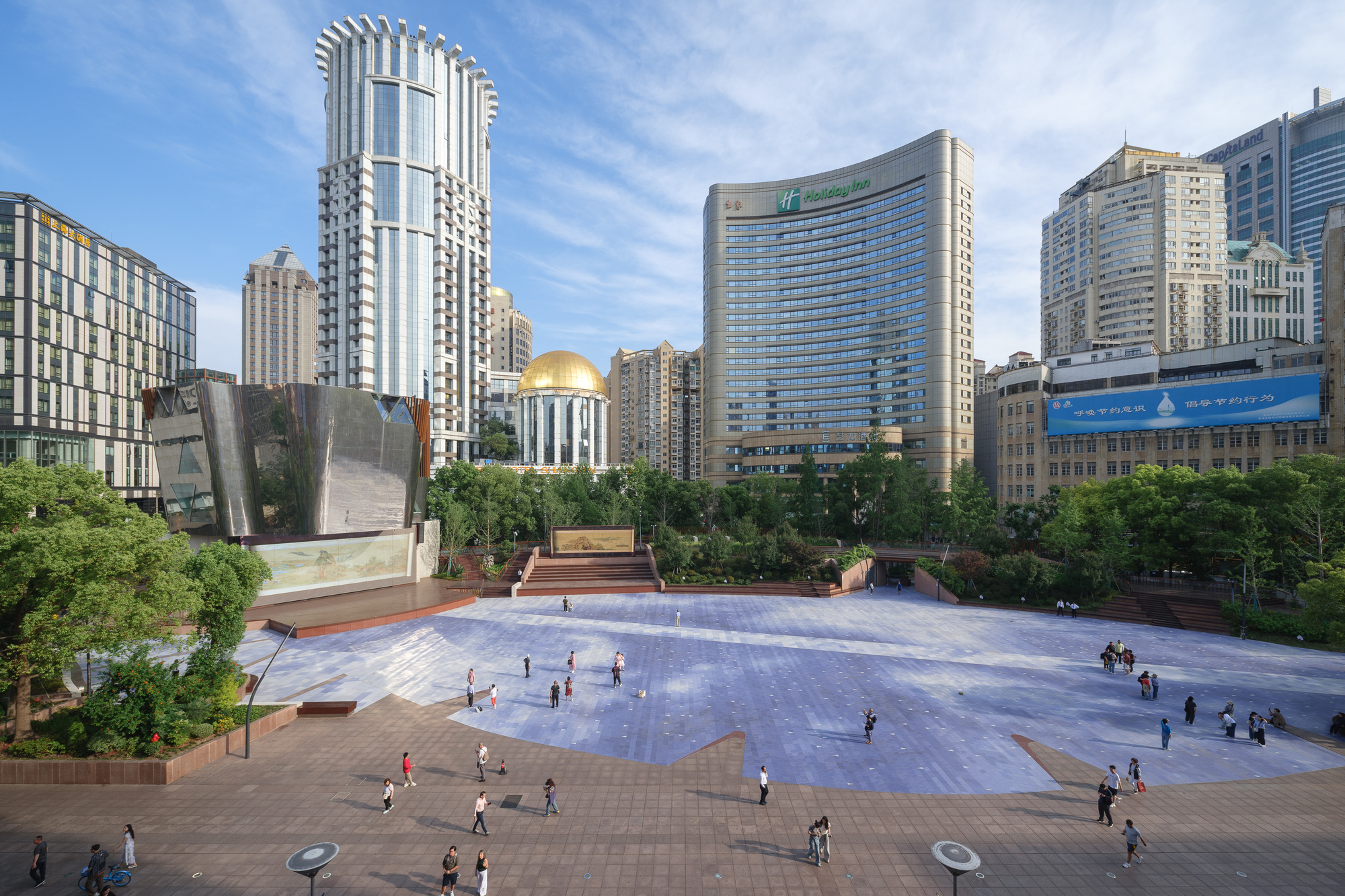Wide view of a plaza with a pale stone-patterned paving field and pedestrians crossing under a blue sky with thin clouds