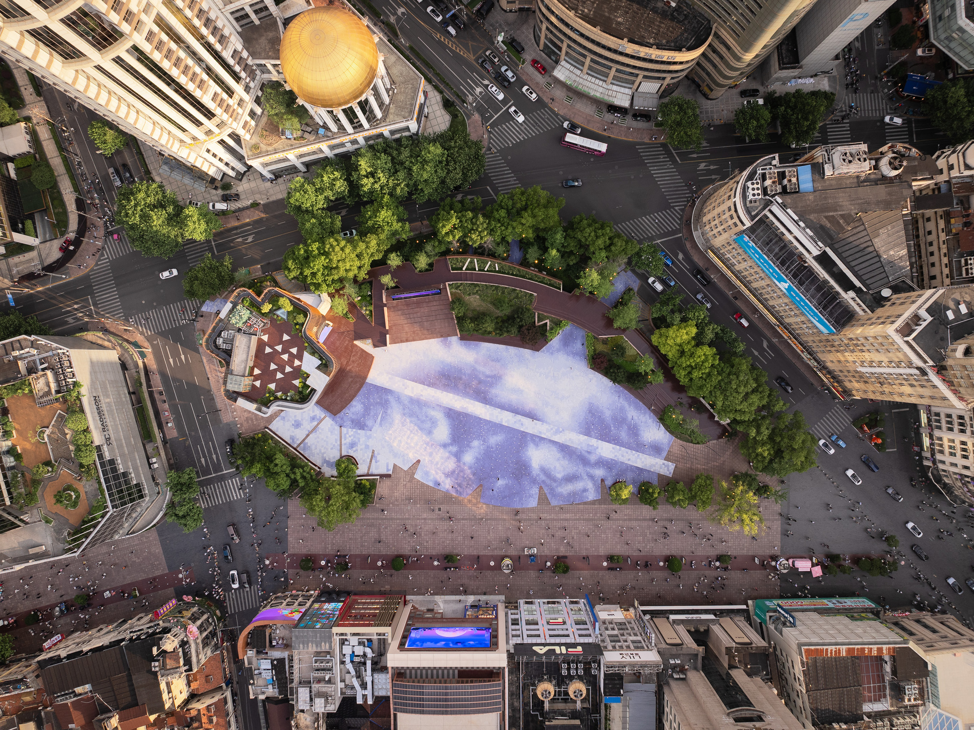 Top-down drone view of the plaza's angular paving, tree groves and rooftop terrace, framed by busy streets at dusk