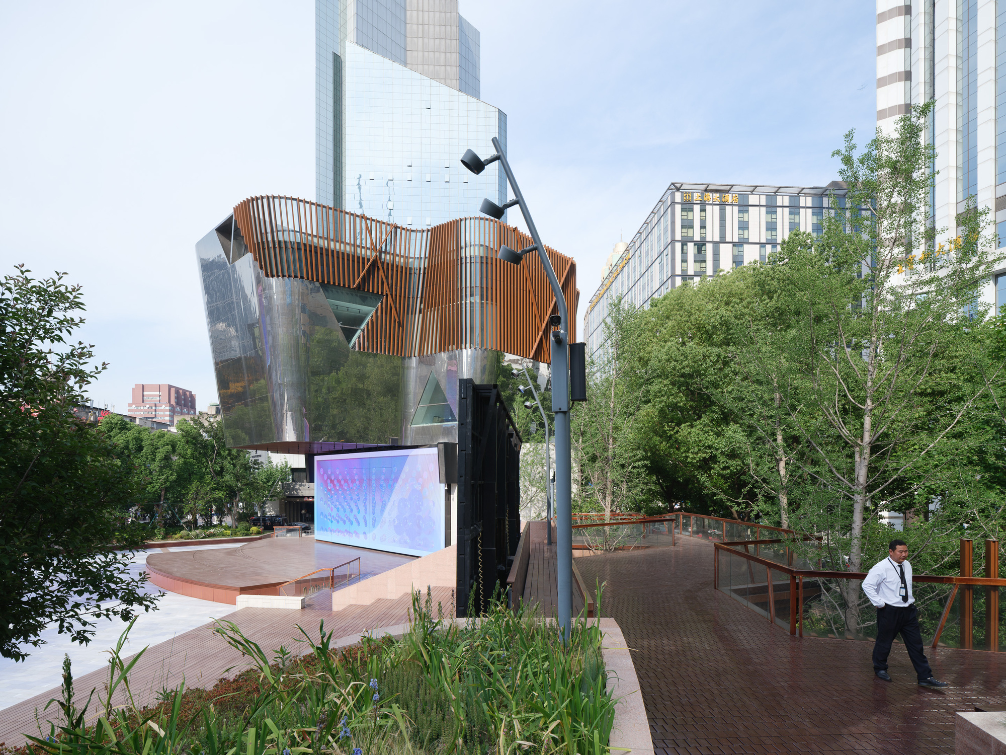 Timber-clad upper volume and LED screen beside a raised walkway of dark wood decking, with a man walking among birch trees