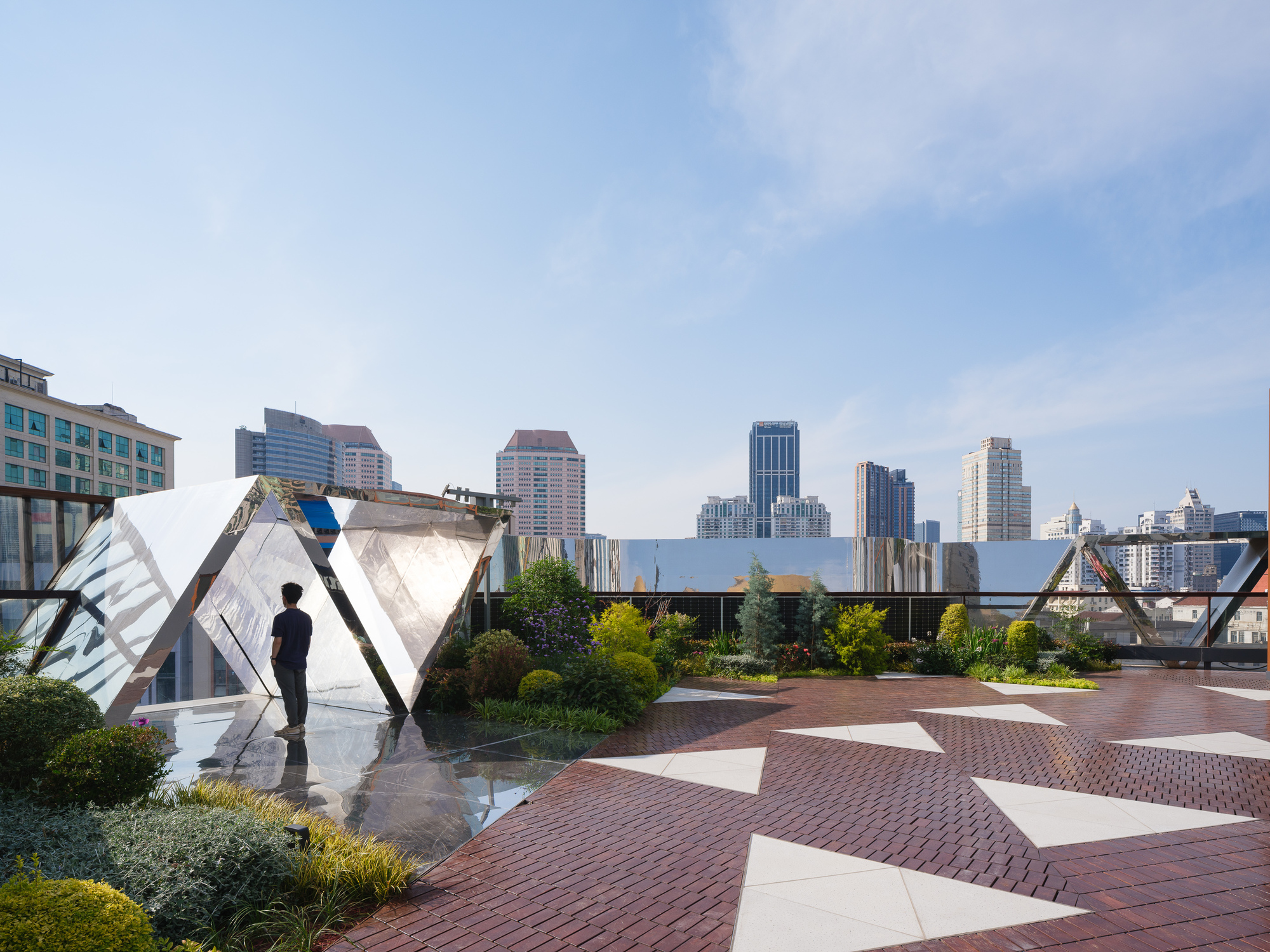 Rooftop garden terrace with faceted mirrored steel canopies, triangular white inlays in red brick paving, and a person looking out at the skyline