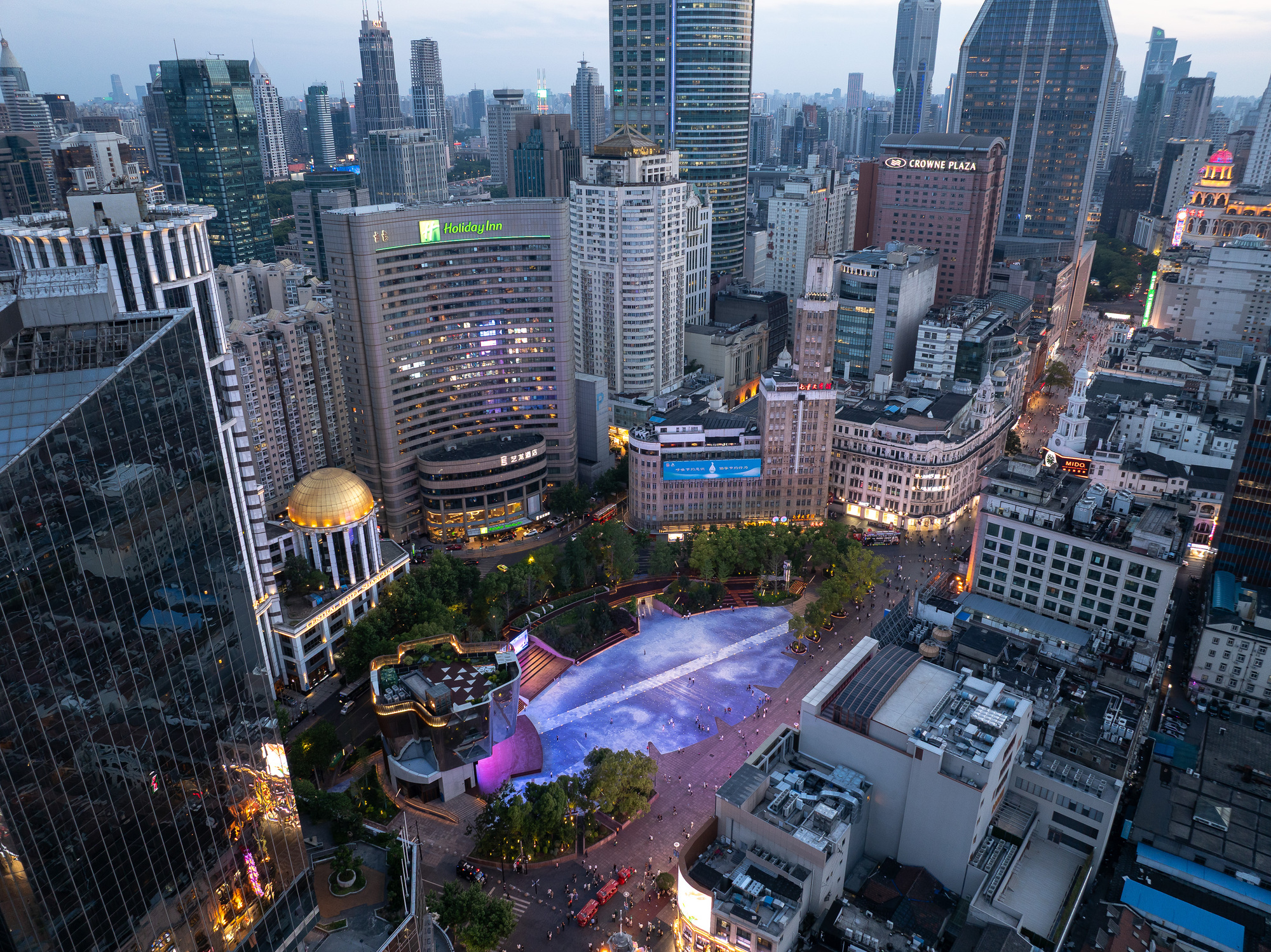 Aerial view at dusk of a sunken plaza with a pale blue paved surface surrounded by towers and lit hotel facades