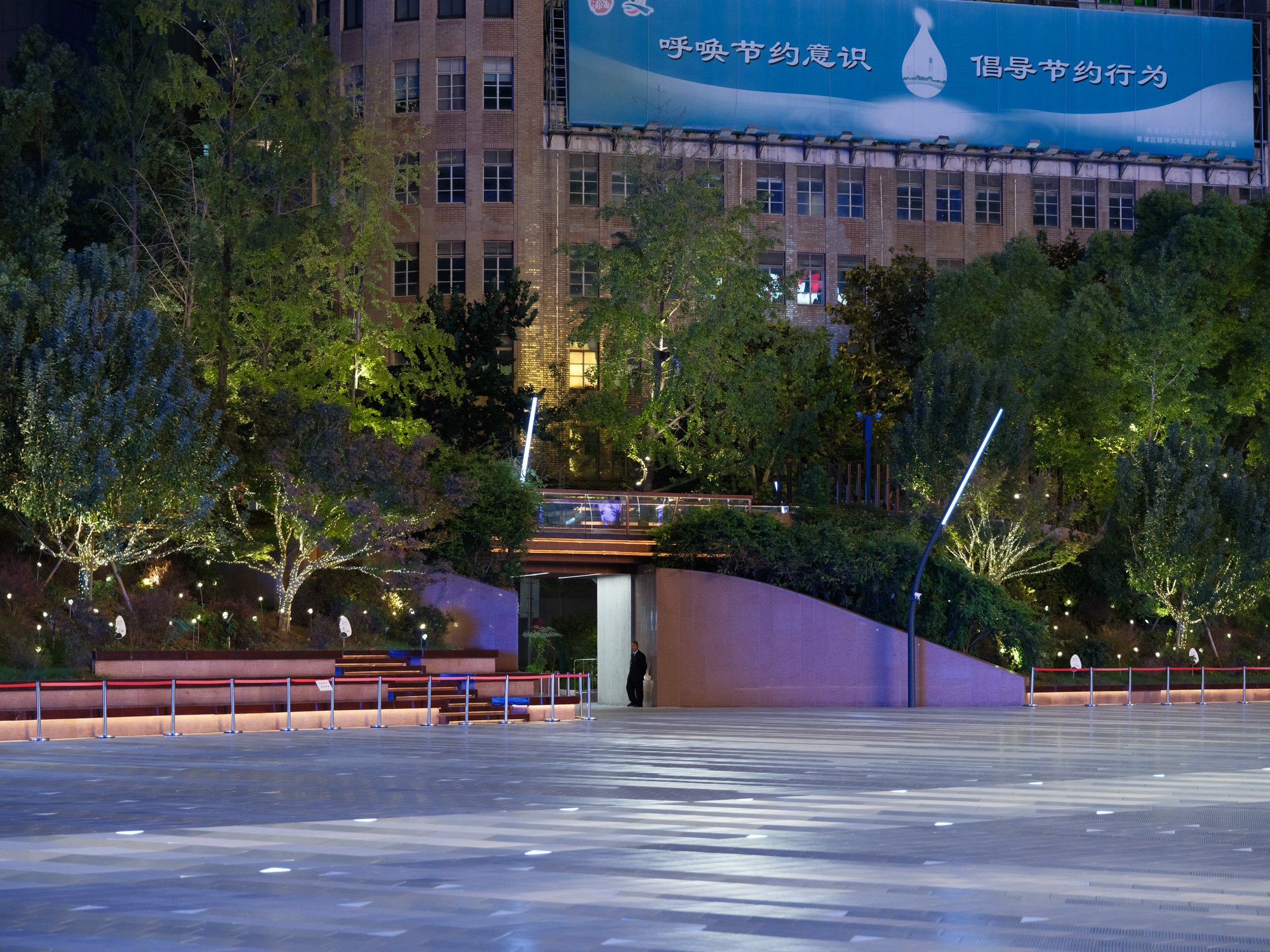 Night view of a paved plaza edged by planted terraces, a curved pink wall and a lone figure at a stair entrance