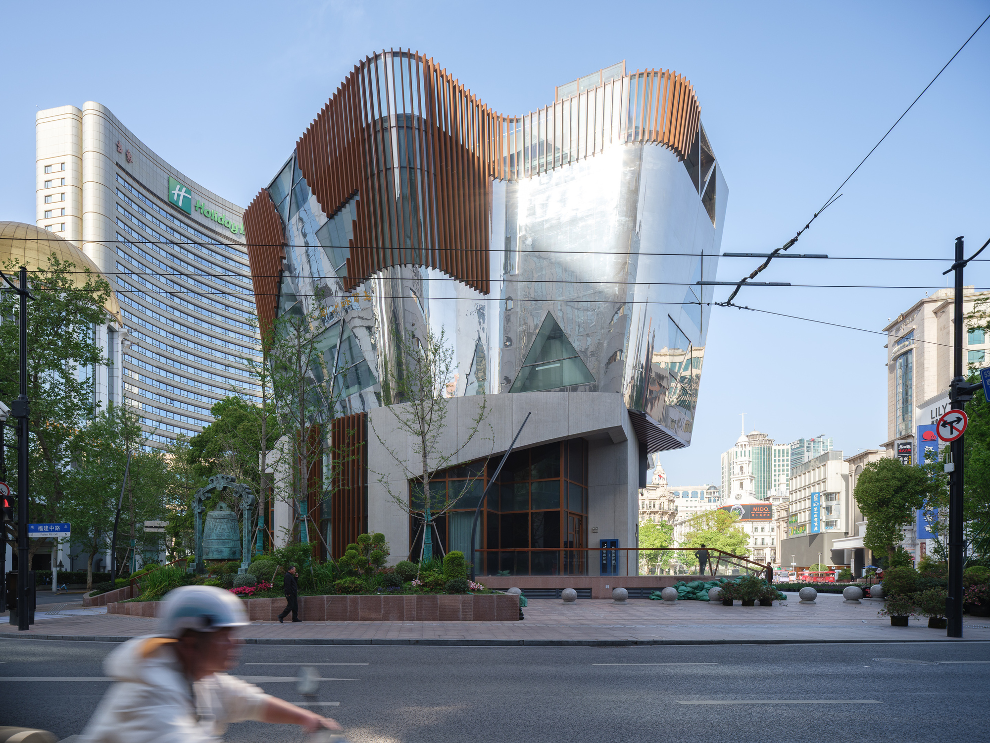 Street corner view of a faceted mirror-steel pavilion with timber fins above a planted base, a scooter rider blurring past