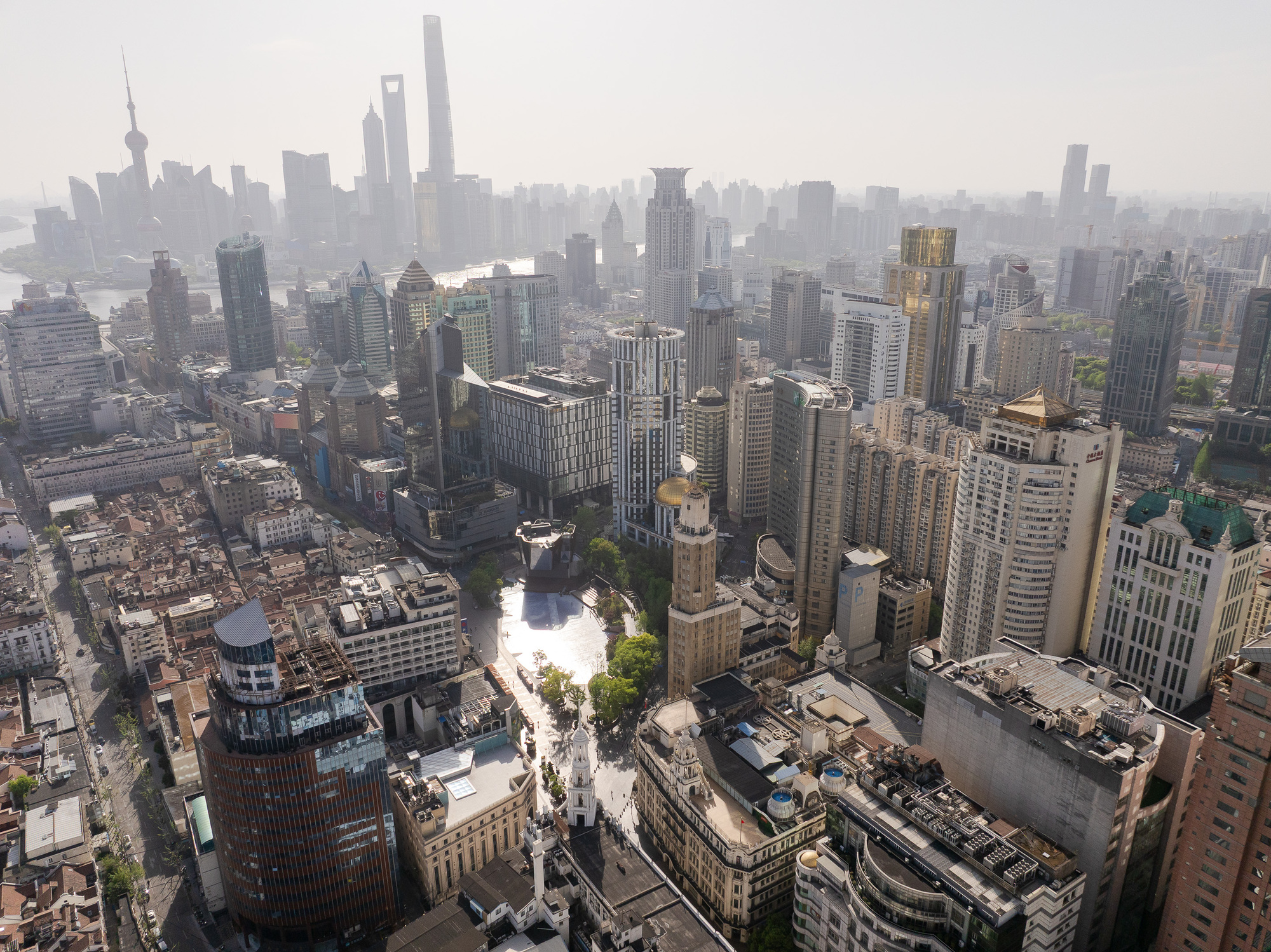 High aerial view of the plaza reflecting sunlight among dense towers and older low-rise blocks, with a hazy skyline behind