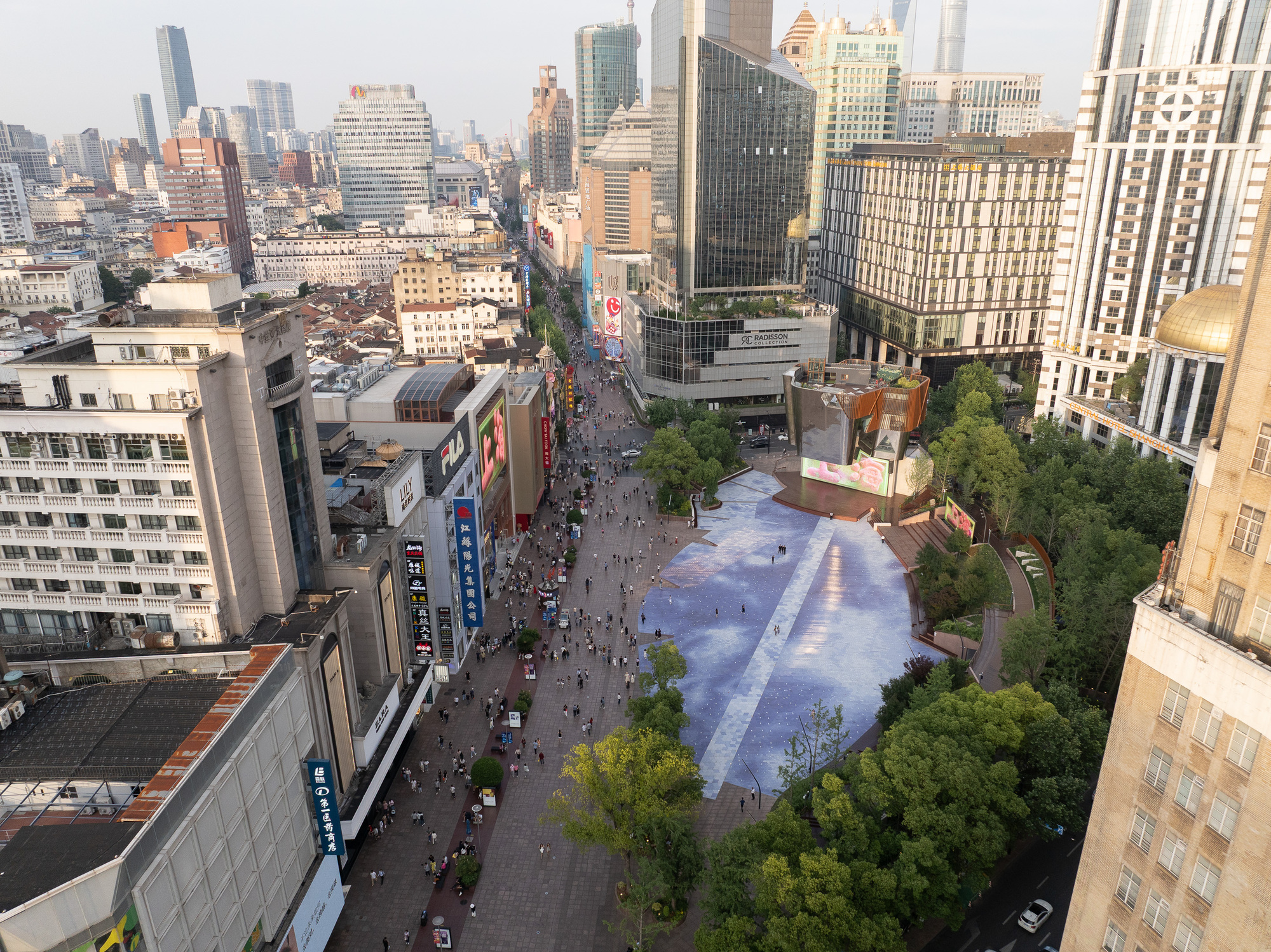 Aerial view of a crowded pedestrian street meeting the plaza, with its pale paving pattern and mirrored pavilion among trees in late sun