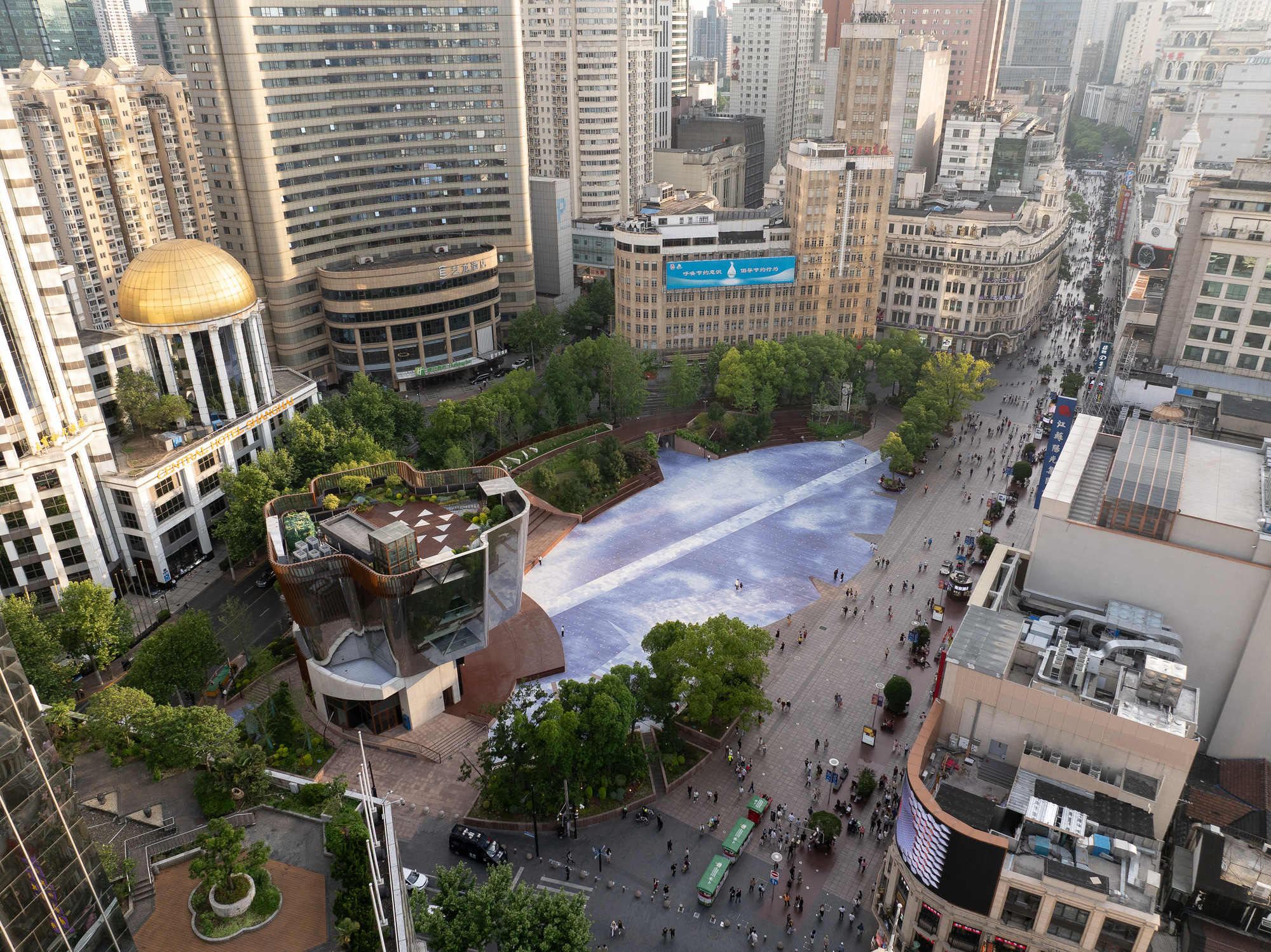 Oblique aerial view of the blue paved plaza, faceted glass pavilion and tree-lined paths among towers in afternoon sun