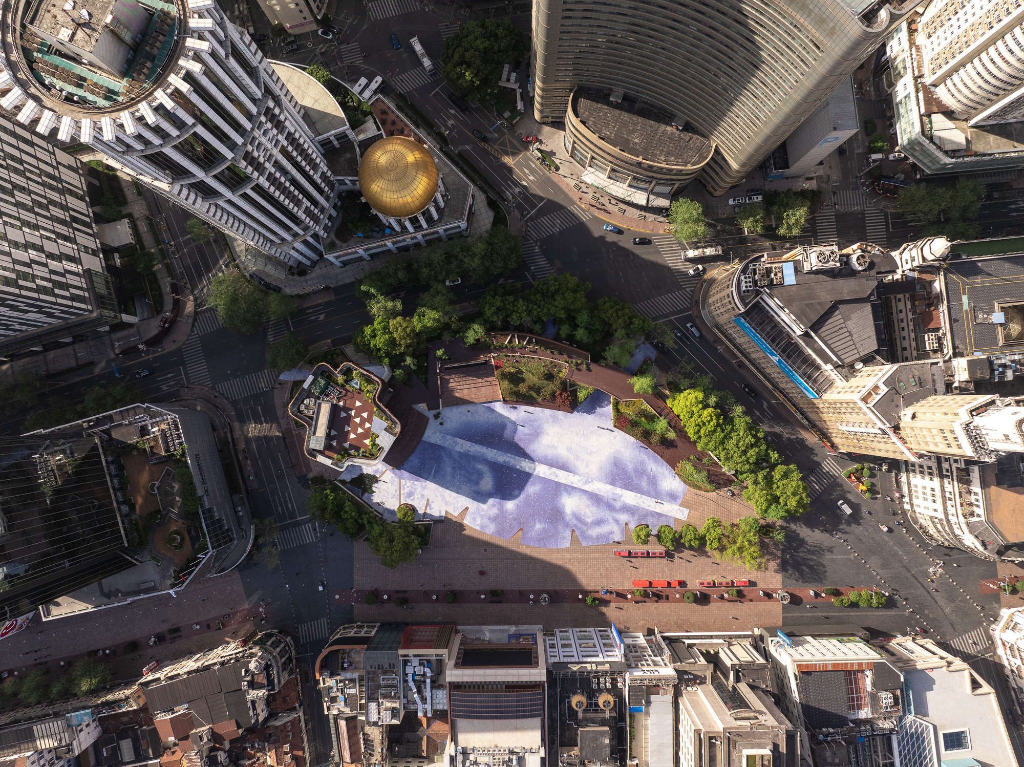 Top-down aerial view of the blue patterned plaza, gold dome and planted edges, with long tower shadows across the ground