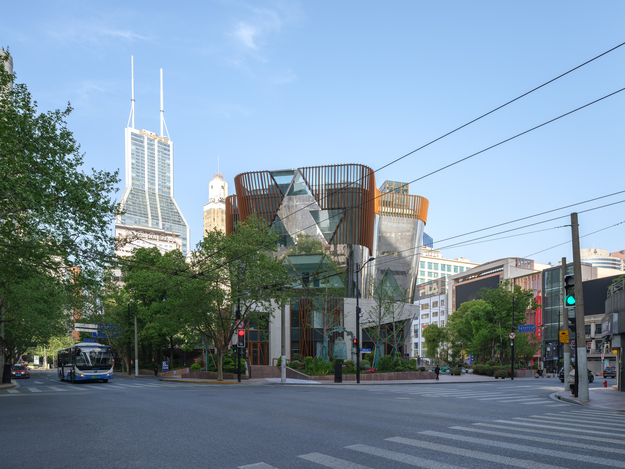 Street corner view of the faceted glass pavilion with copper-toned vertical fins, framed by plane trees under a clear blue sky