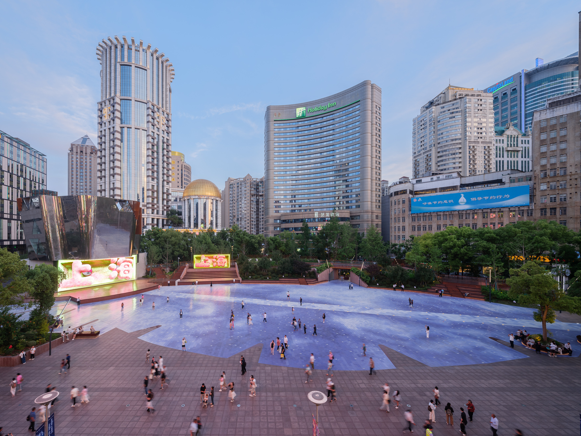 Wide view of the blue paved plaza with blurred pedestrians crossing, a mirrored volume at left and a gold dome at dusk