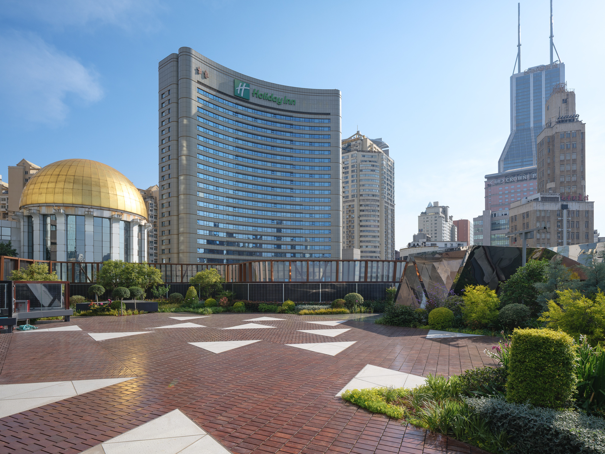 Rooftop terrace with brick paving, white triangular inlays and clipped shrubs, facing a curved hotel tower and gold dome