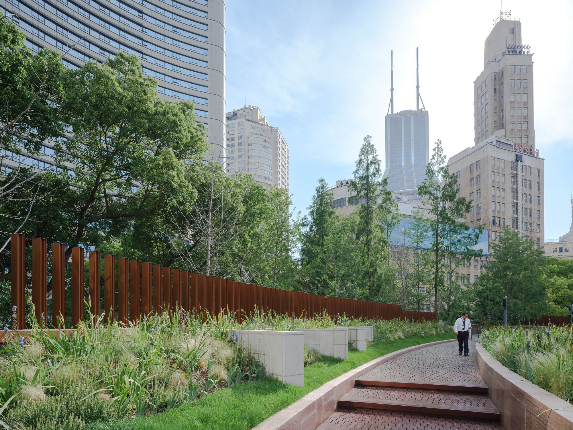 Curving brick ramp with stepped edges beside a weathering steel fence and grasses, with a man in a white shirt walking