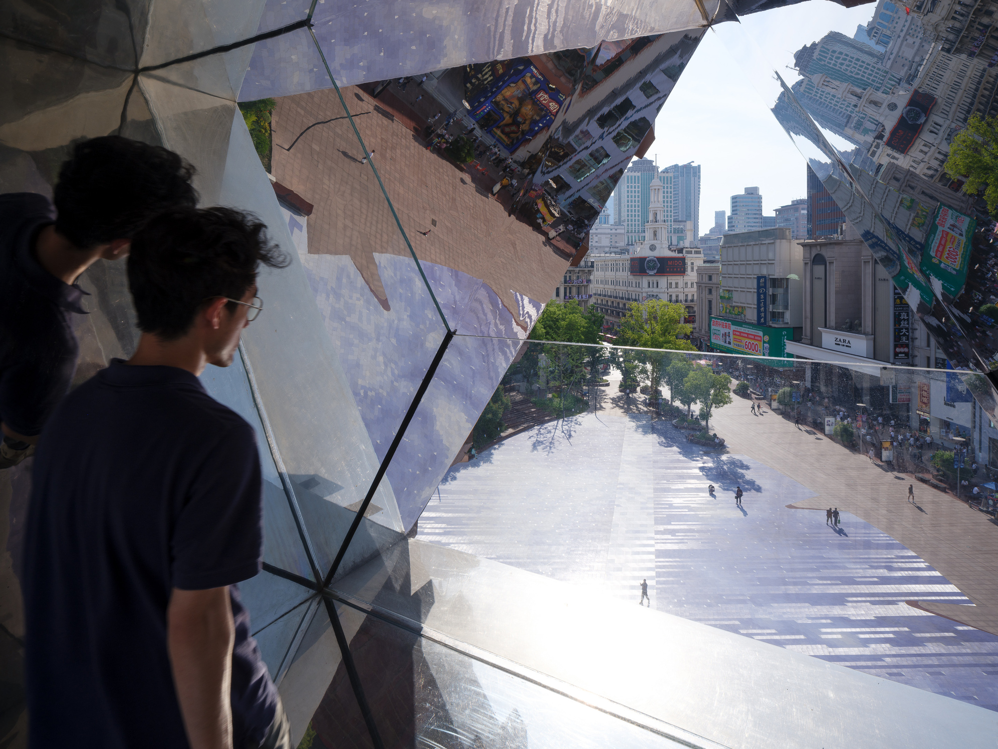 Two people look out through a faceted mirrored glass enclosure toward a wide paved plaza and distant towers in afternoon sun