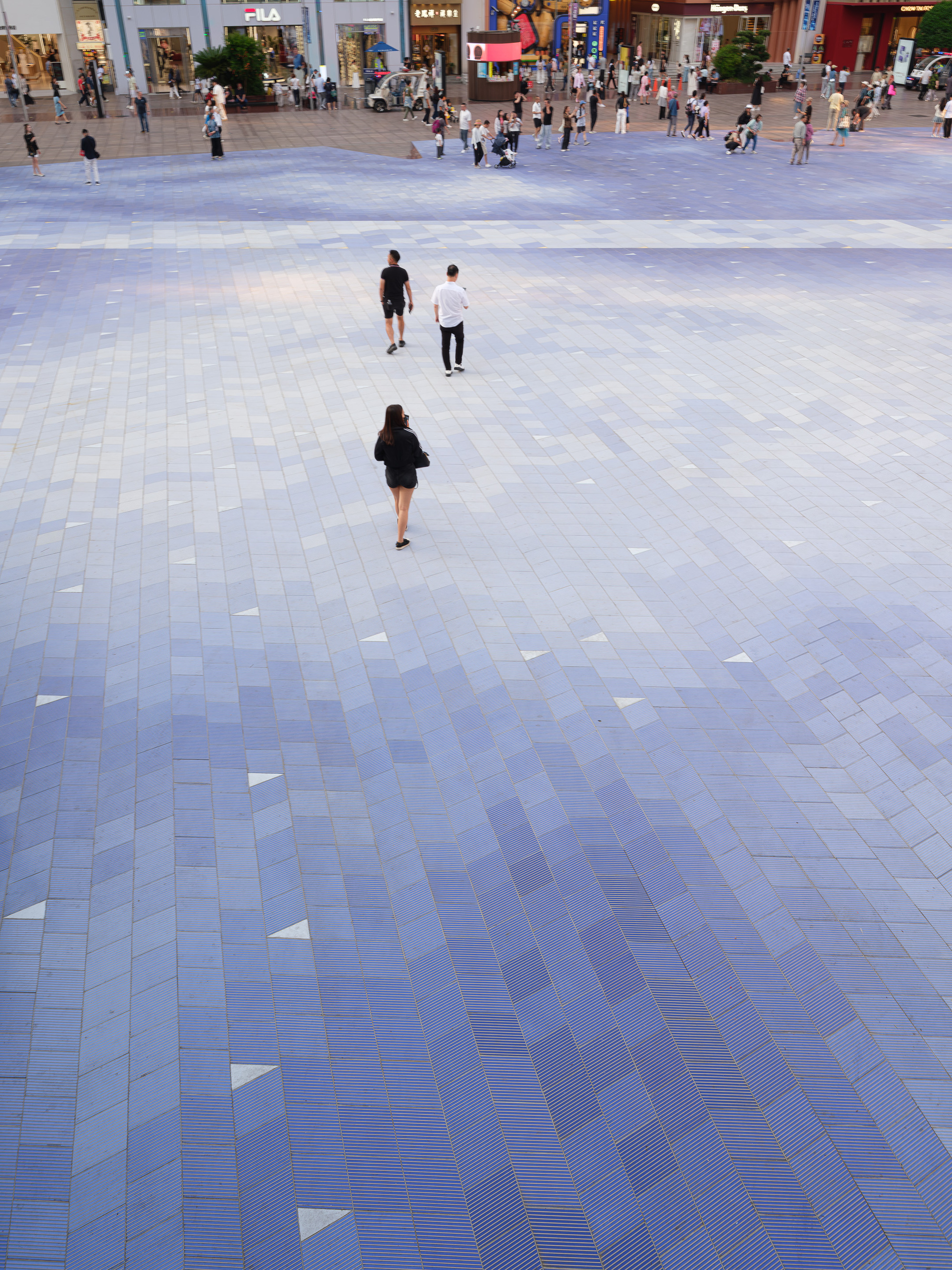 High view of the blue gradient paving across a plaza, with three pedestrians walking toward storefronts at dusk